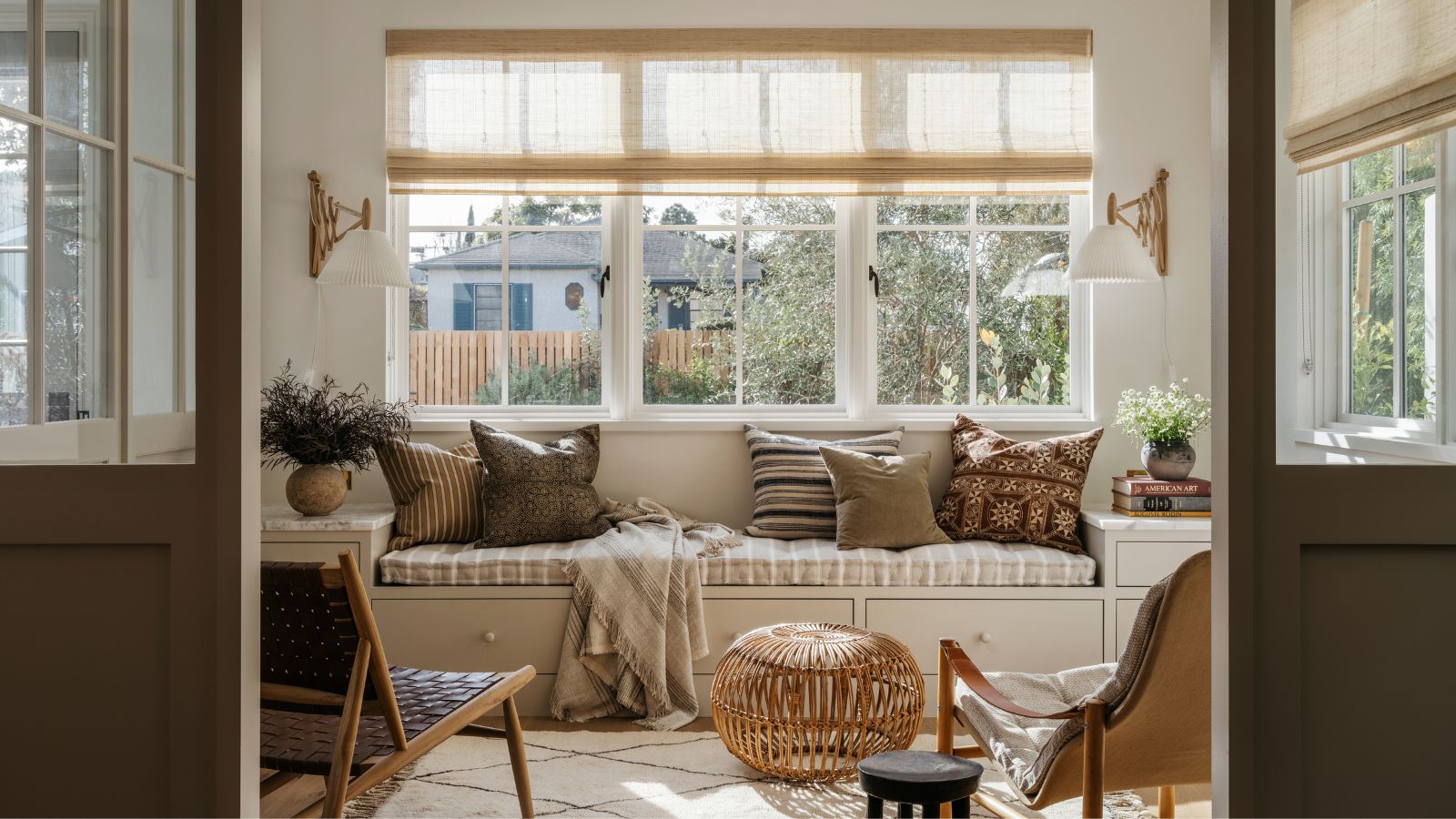 Lounge im Küstenstil mit eingebauter Bank, dekoriert mit neutralen Kissen, einem Berber-Teppich, Mid-Century-Loungesesseln aus Holz und einem Rattan-Ottomane 