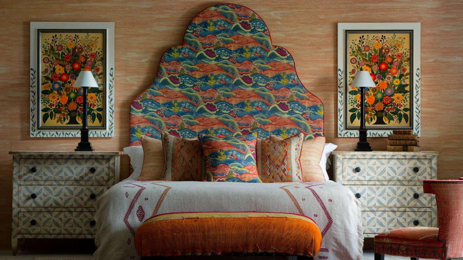 Ein farbenfrohes Schlafzimmer mit einem großen Bett mit übergroßem gemustertem Kopfteil, weißer Bettwäsche, verschiedenen bunten Kissen und einem orangefarbenen Ottoman am Ende des Bettes. Auf beiden Seiten befinden sich weiße Kommoden mit grünen geometrischen Mustern und darüber braune Lampen mit kleinen weißen Lampenschirmen. Hinter beiden Lampen befinden sich große Blumenkunstwerke in weißen Rahmen, und die Wand ist in einem blass gebrannten Orange mit weißen Details tapeziert.