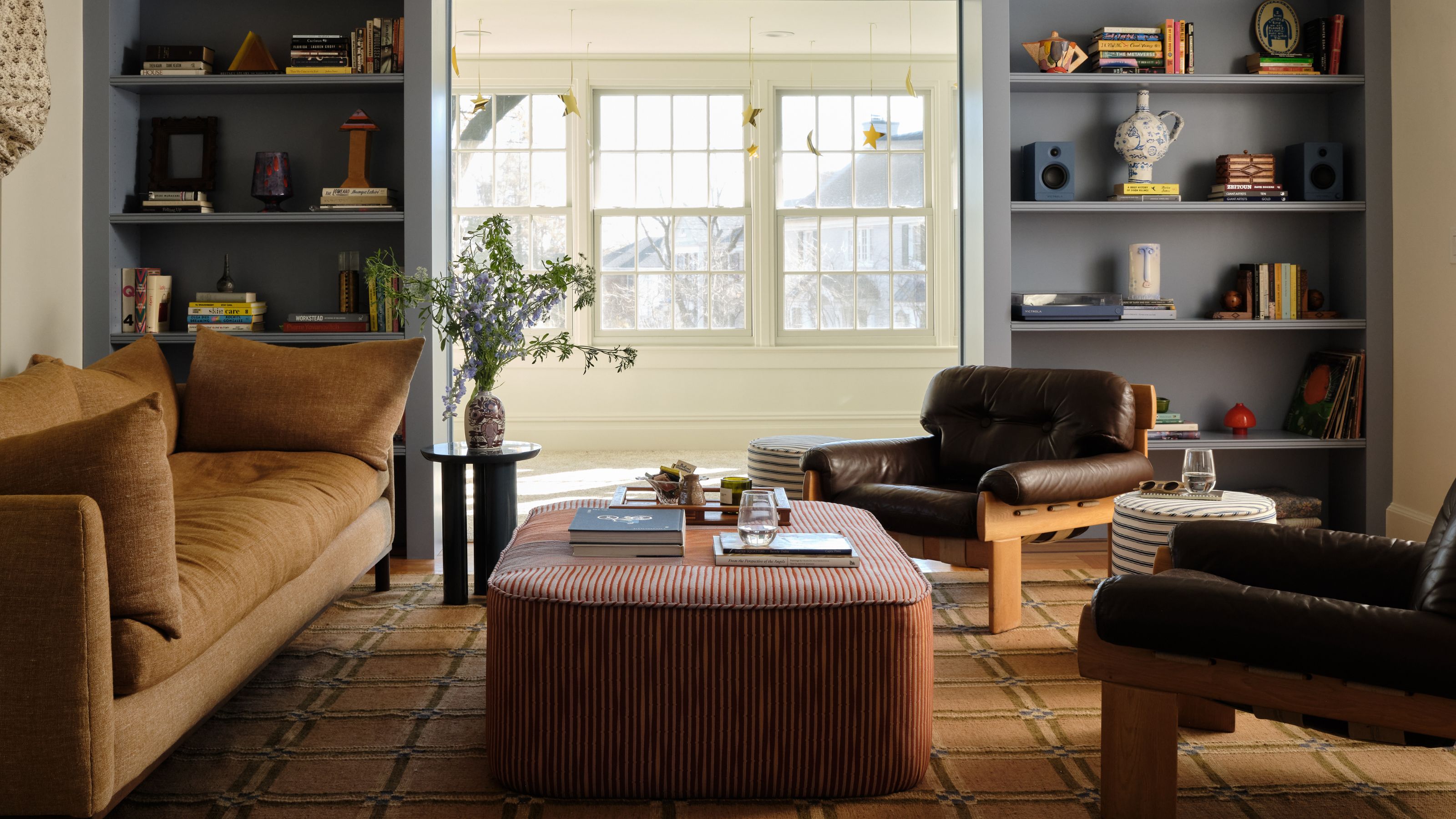 Ein kleines Wohnzimmer in New Jersey mit großen Fenstern, maßgefertigten blauen Bücherregalen, einem Teppich aus Westulme und einem Sofa im Vintage-Stil der 1970er Jahre sowie Ledersesseln