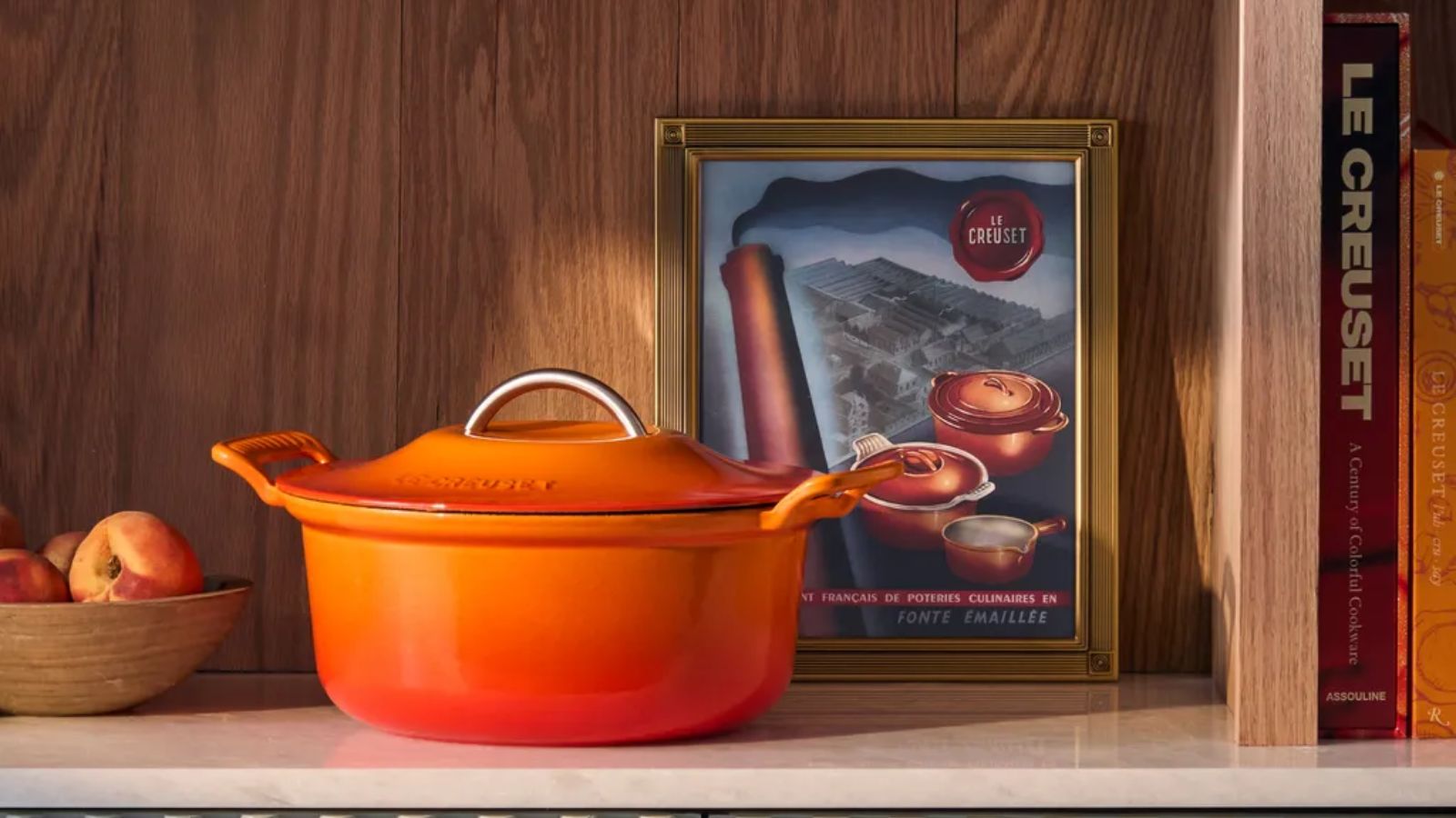 Ein orangefarbener Dutch Oven auf einem Holzregal, daneben eine gerahmte Illustration der Le Creuset Modern Heritage Pfannenkollektion und eine Holzschale mit Äpfeln. 