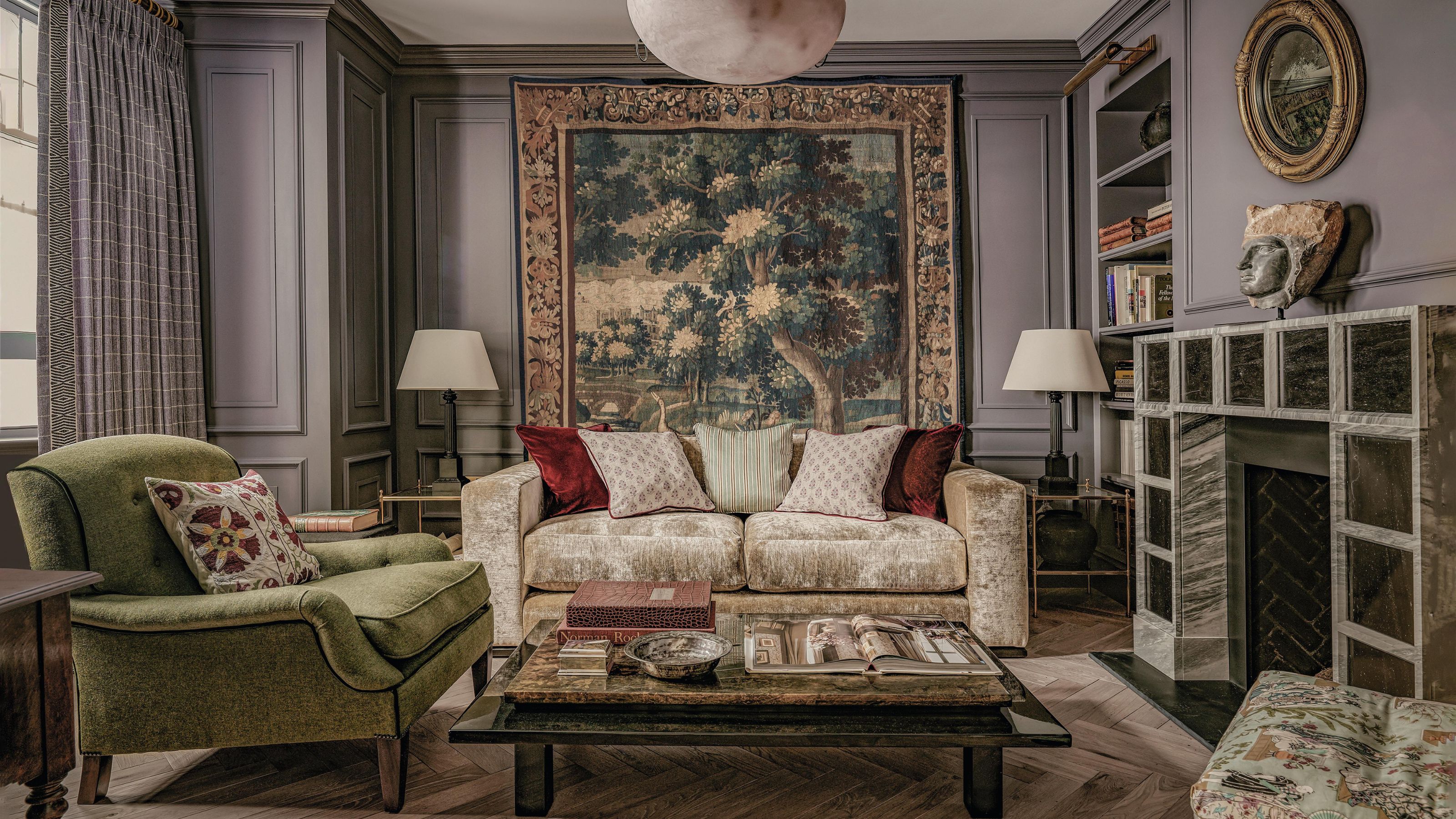 ein malvenfarbenes Wohnzimmer mit einem großen Vintage-Wandteppich, Wandverkleidung, einem einzigartigen quadratischen Kamin, einem cremefarbenen Samtsofa und einem grünen Sessel