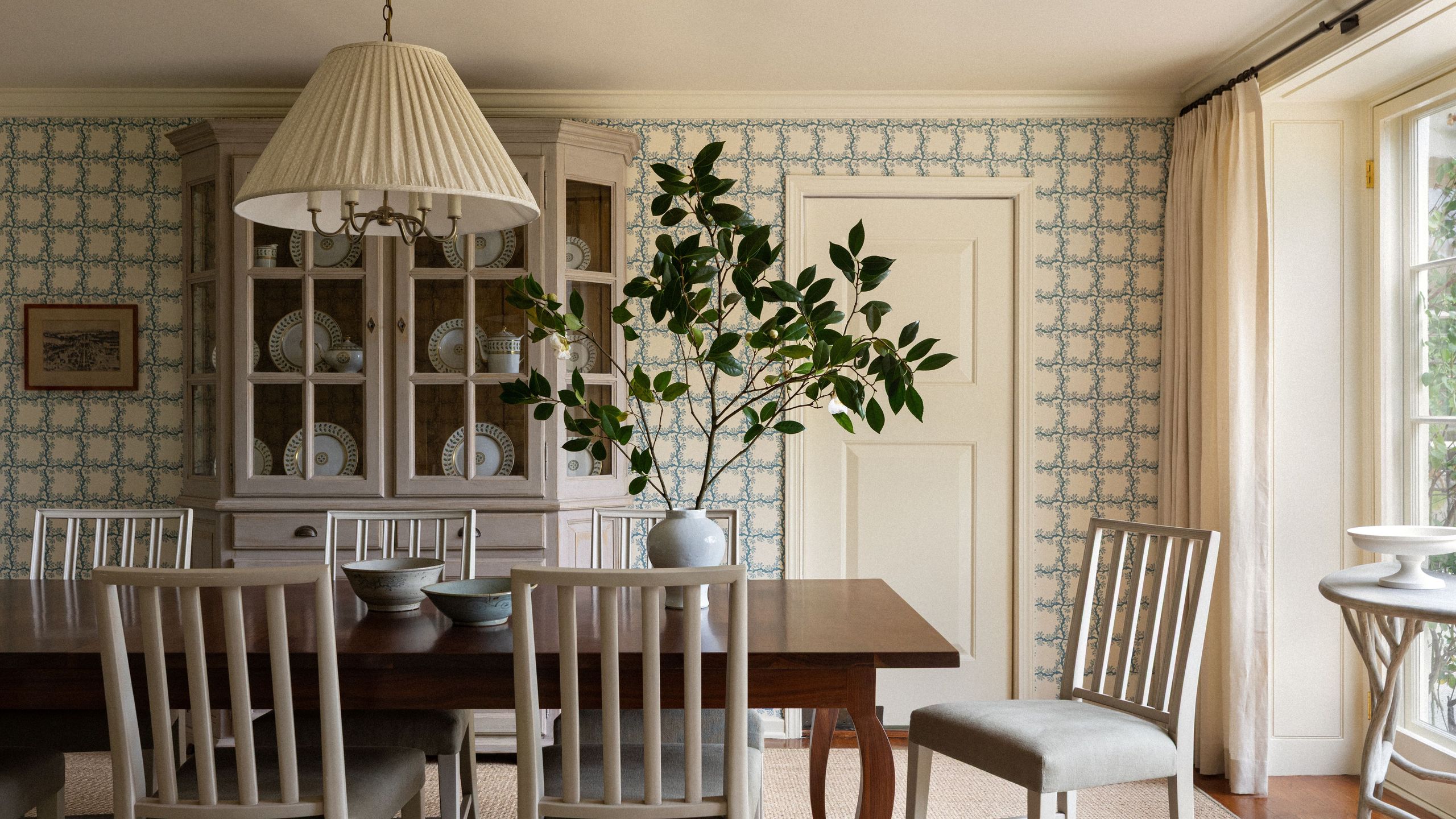 Sun-drenched dining room featuring blue-and-white rose-motif wallpaper, airy white dining chairs, a pleated pendant light, and a large potted plant