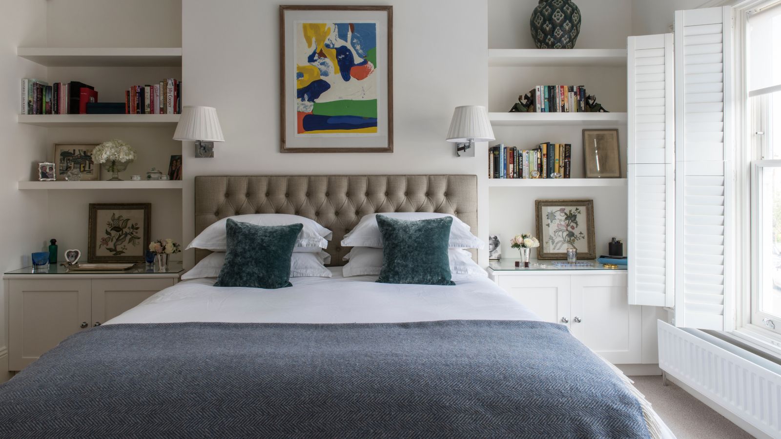 Ein Doppelzimmer mit weißer Bettwäsche mit blauen Akzenten und großen Fenstern auf der rechten Seite mit offenen weißen Fensterläden. Auf beiden Seiten des Bettes befinden sich Regale mit Büchern, Vasen und gerahmten Bildern, und über dem Bett befinden sich zwei weiße Wandleuchten und ein farbenfrohes Kunstwerk.