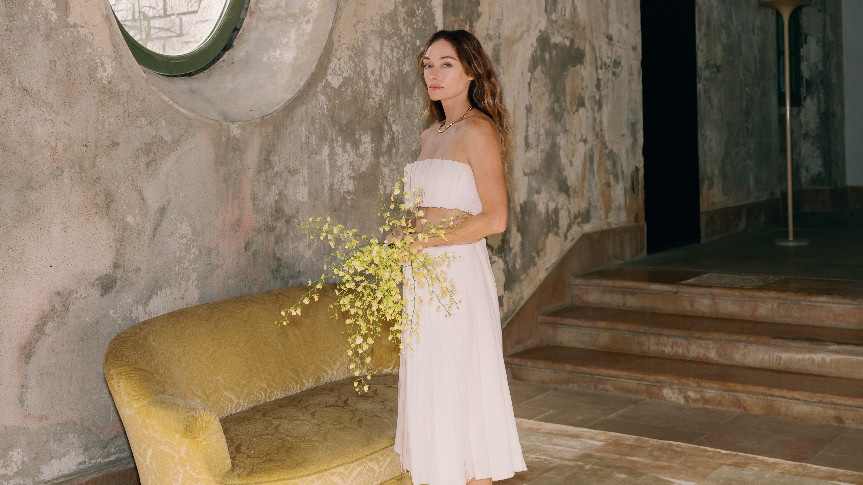 Kelly Wearstler steht in einem brutalistischen Raum und hält einen Strauß gelber Blumen