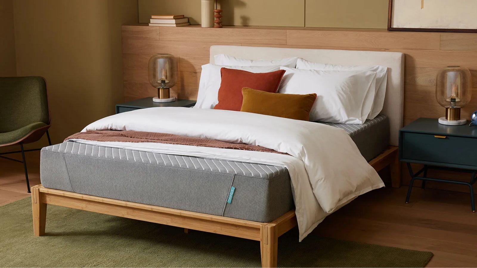 Eine graue Siena-Matratze auf einem weißen Bett abgebildet. mit Holzsockel, vor einer holzvertäfelten Wand. Der Bodenbelag besteht aus dem gleichen hellen Holz mit einem grünen Teppich, und zu beiden Seiten des Bettes stehen passende blaue Nachttische mit Lampen. Links daneben steht auch ein grüner Stuhl. Oberhalb der Holzvertäfelung ist die Wand hellgrün.