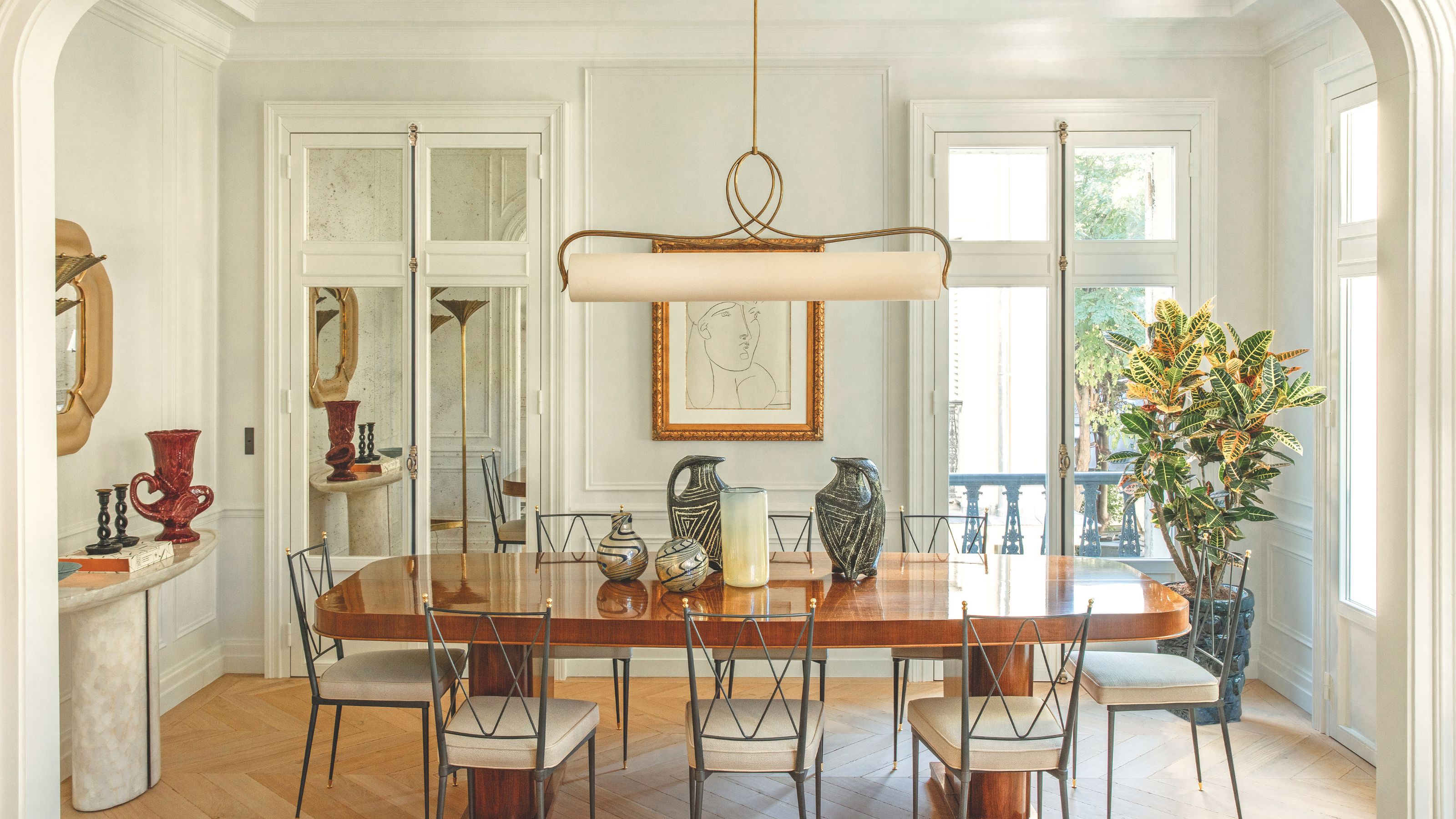 Ein elegantes, sonnendurchflutetes Esszimmer mit einem polierten Holztisch, schlanken schwarzen Metallstühlen und einer einzigartigen horizontalen Pendelleuchte. Der Raum ist mit Keramikvasen und einer großen gerahmten Skizze an einer getäfelten Wand dekoriert.