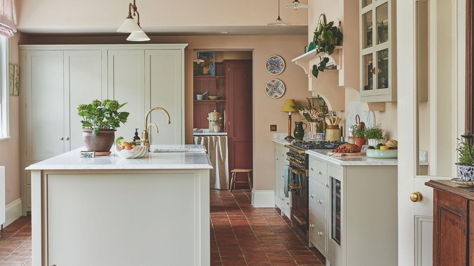 Eine rustikale Küche im Landhausstil mit hell gestrichenen Schränken und Kochinsel sowie weißen Arbeitsplatten. Rosa gestrichene Wände. 