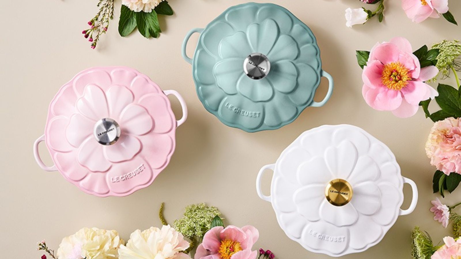 Drei pastellfarbene Le Creuset Dutch Ovens mit Blumenmuster auf einem cremefarbenen Hintergrund, umgeben von rosa und weißen Blumen