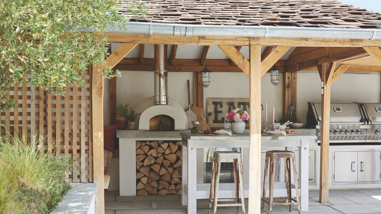 An outdoor kitchen with stainless steel countertops under a wooden trellis and tiled roof. Complete with a pizza oven