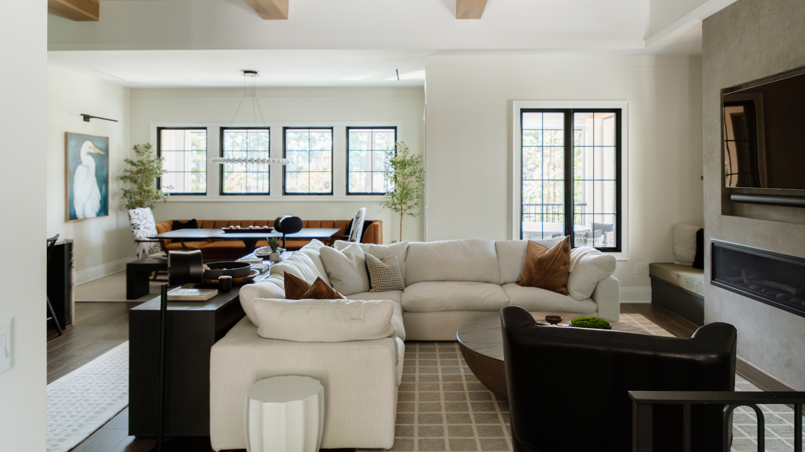 Ein einladendes Wohnzimmer mit einer großen cremefarbenen Sitzecke, einem Ledersessel, einem gemusterten Teppich, Kissen, einem dunklen Konsolentisch hinter der Couch mit Deko und einem in die Wand eingebauten Fernseher über einem Kamin 