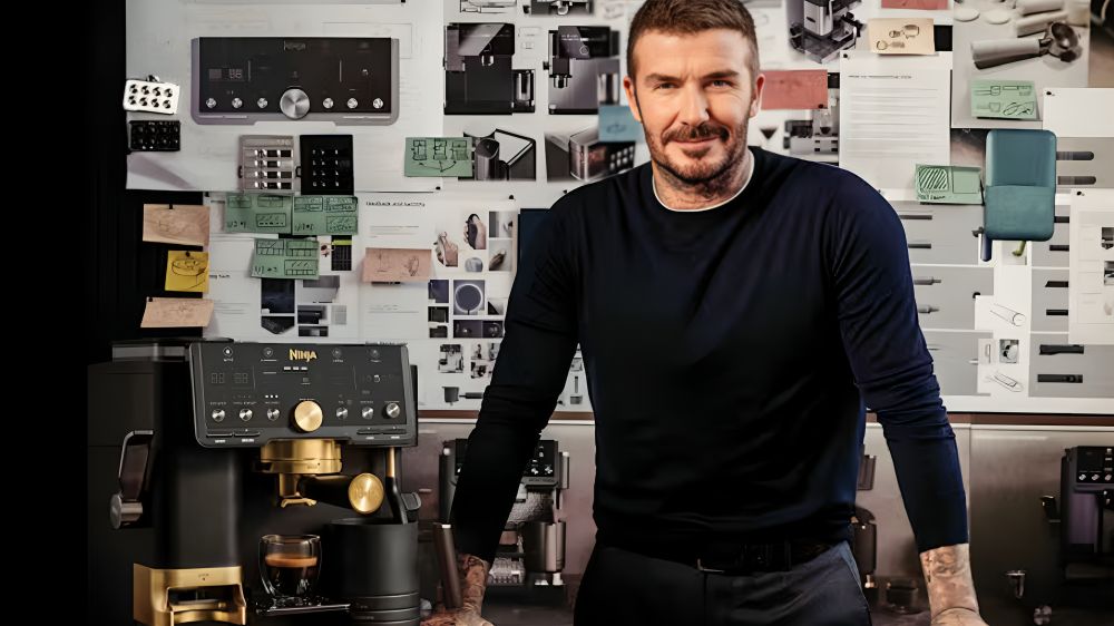 David Beckham stands in front of a large vision board, beside a black and gold coffee maker.