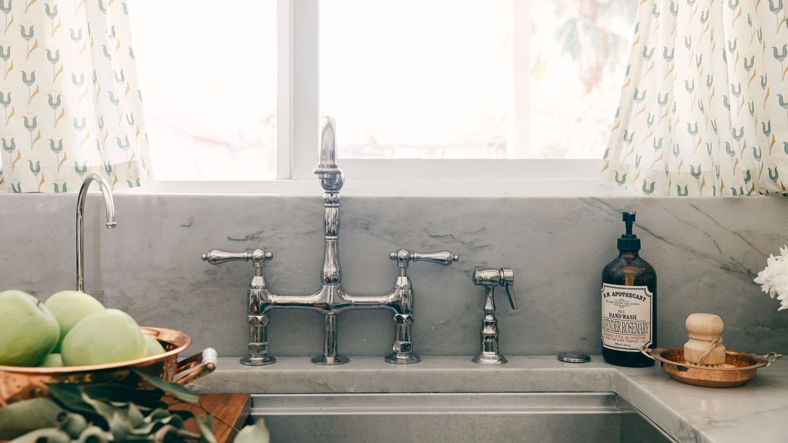 Kitchen faucet in front of bright, sunny window. A fruit bowl is pictured to the left, and soap to the right.