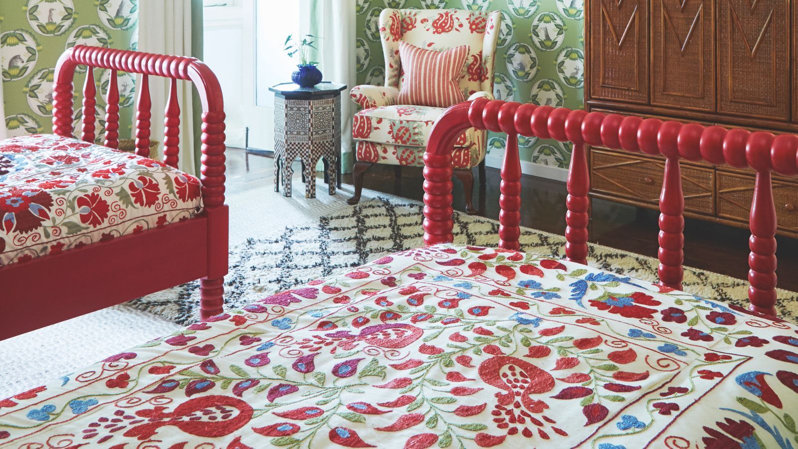 A bright, maximalist twin bedroom with red beds, colorful bedding, patterned rugs, wallpaper, upholstered chair, and side table. There's a large wooden wardrobe, and windows to the back left corner.