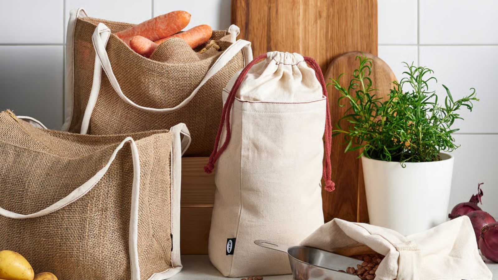 A set of cotton and jute food storage bags on a kitchen counter, beside some wooden cutting boards and a metal plant pot of herbs.