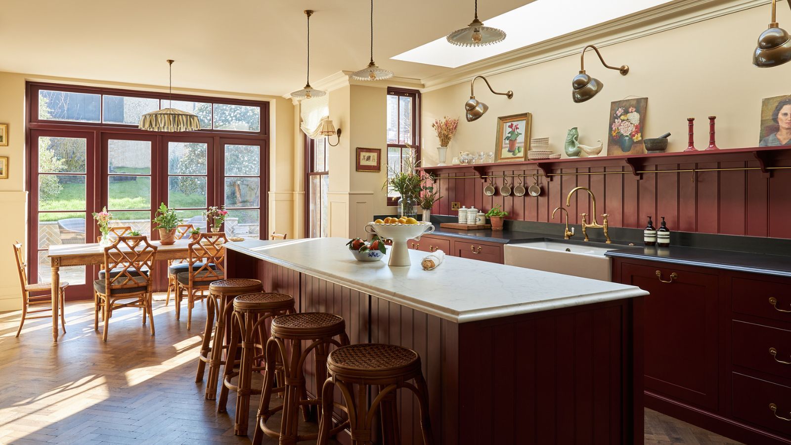 A large kitchen with the cabinets and island painted oxblood red, a paneled backsplash, black countertops on the perimeter cabinets, and a white marble counter on the island.