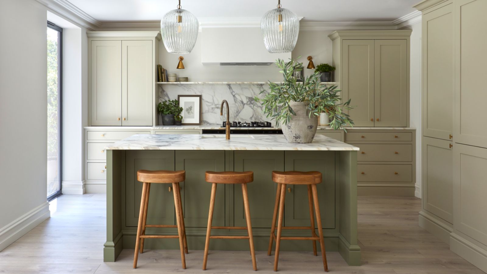 A kitchen with sage green cabinets and a darker green island. A large stone vase with green steps on the island beside a sink, and two glass pendants hanging from the ceiling above.