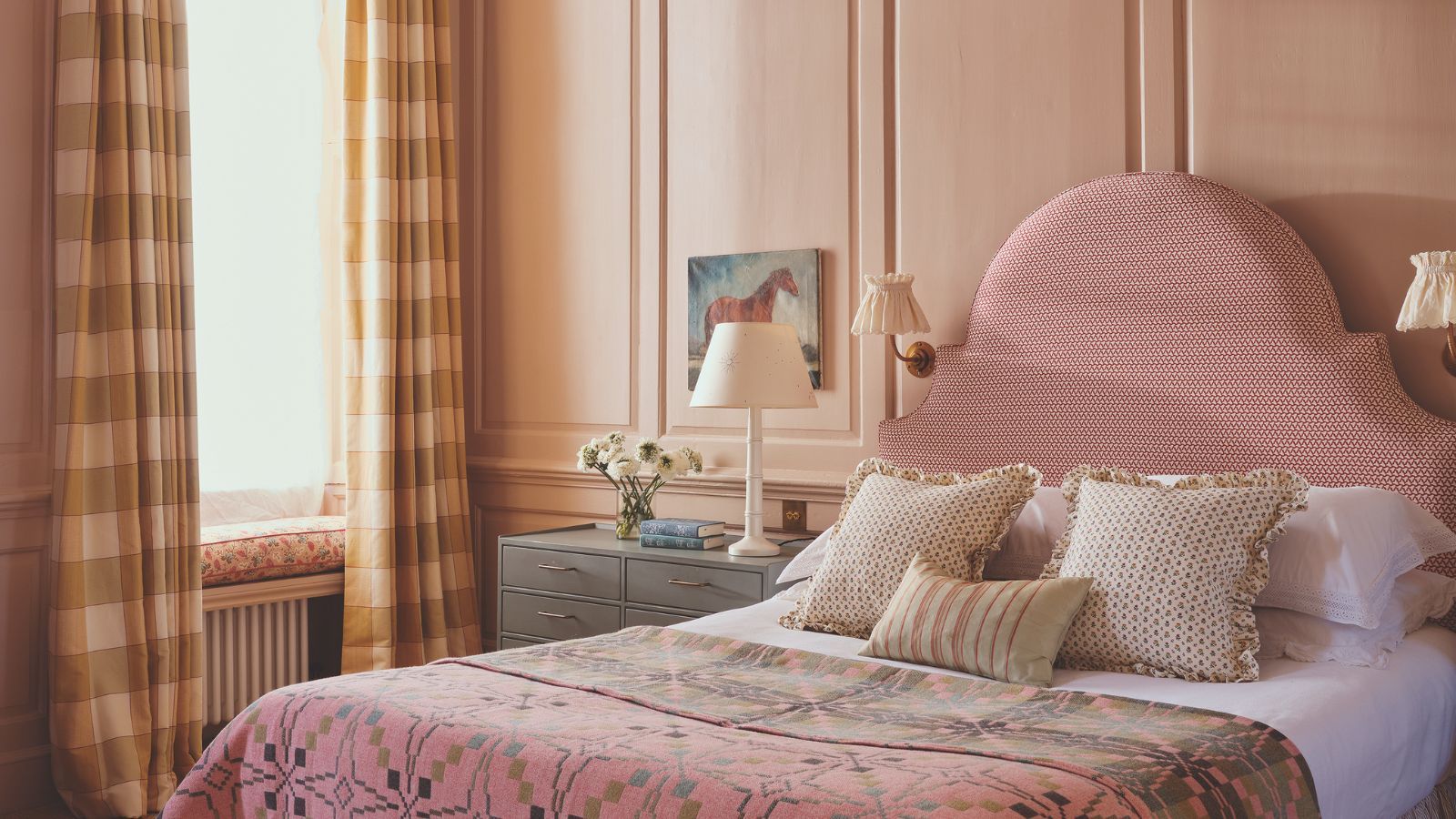 Bedroom with warm pink walls, upholstered pink bed with curved headboard, a pink quilt, striped throw pillows and white bed linen, a nightstand with a horse painting above it and plaid yellow curtains