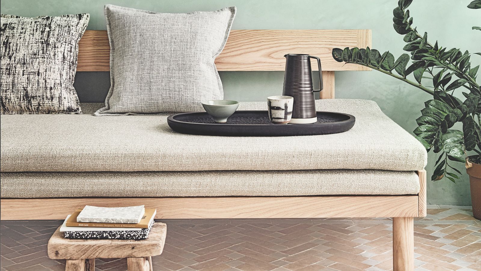 A sage green room with Japanese futon sofa with cushions, tray with teapot and cups, and a wooden stool with books. The tiles are pale terracotta, with a large houseplant to the right.