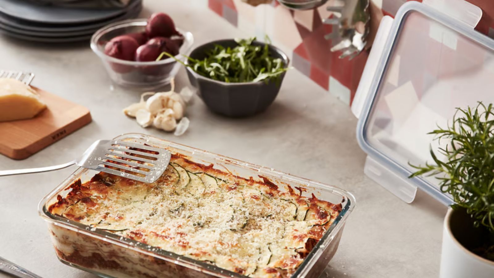 Gemüselasagne im Glasbehälter neben Schüsseln mit Kräutern und Gewürzen