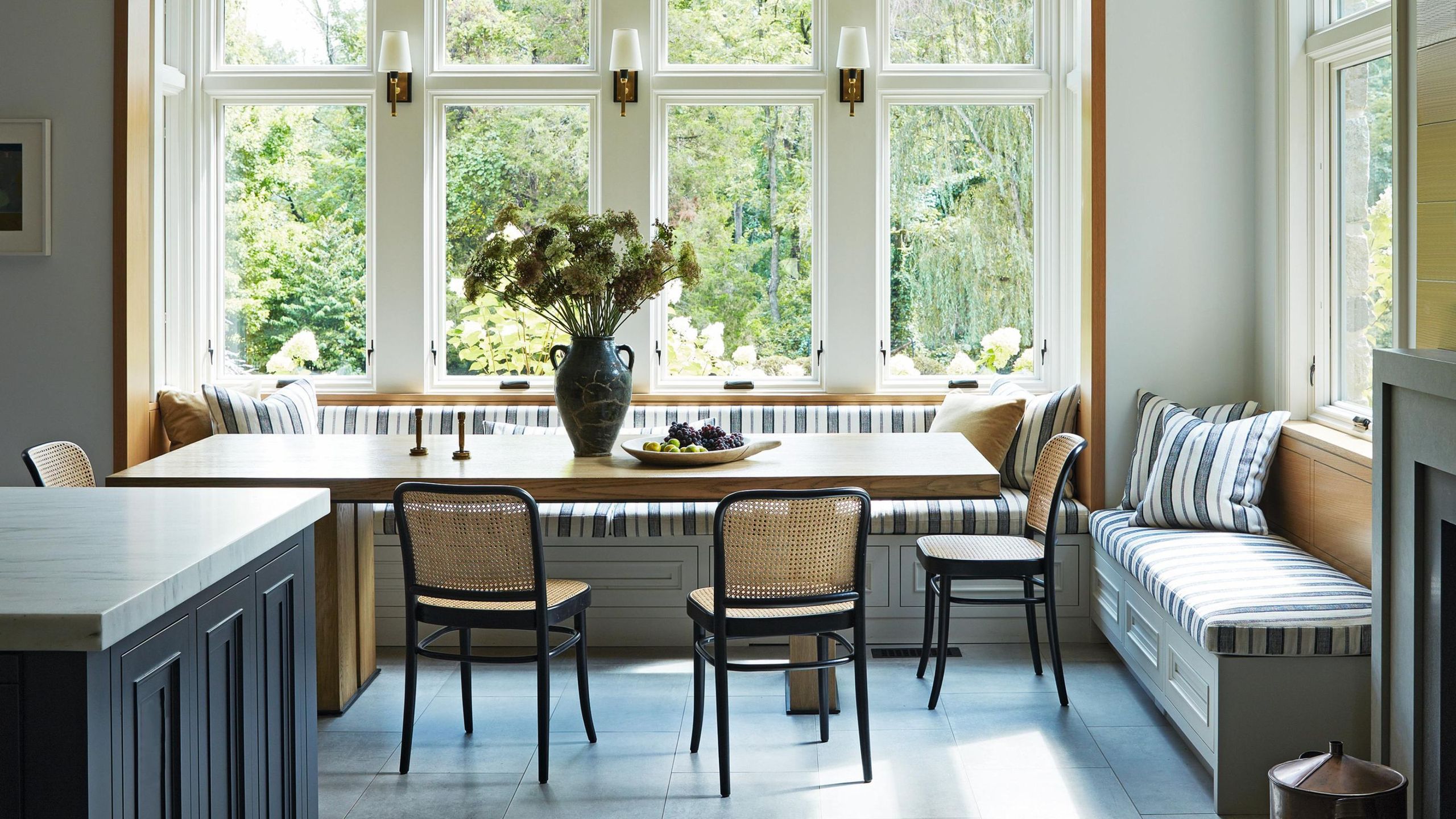 a light and bright kitchen dining area with a striped large banquette, ceiling beams and blue tiled floor