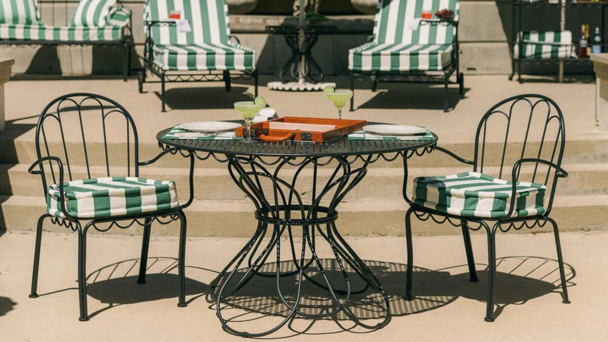 Whimsical iron outdoor bistro table and chairs, pictured in front of a row of loungers with green-and-white striped cushions