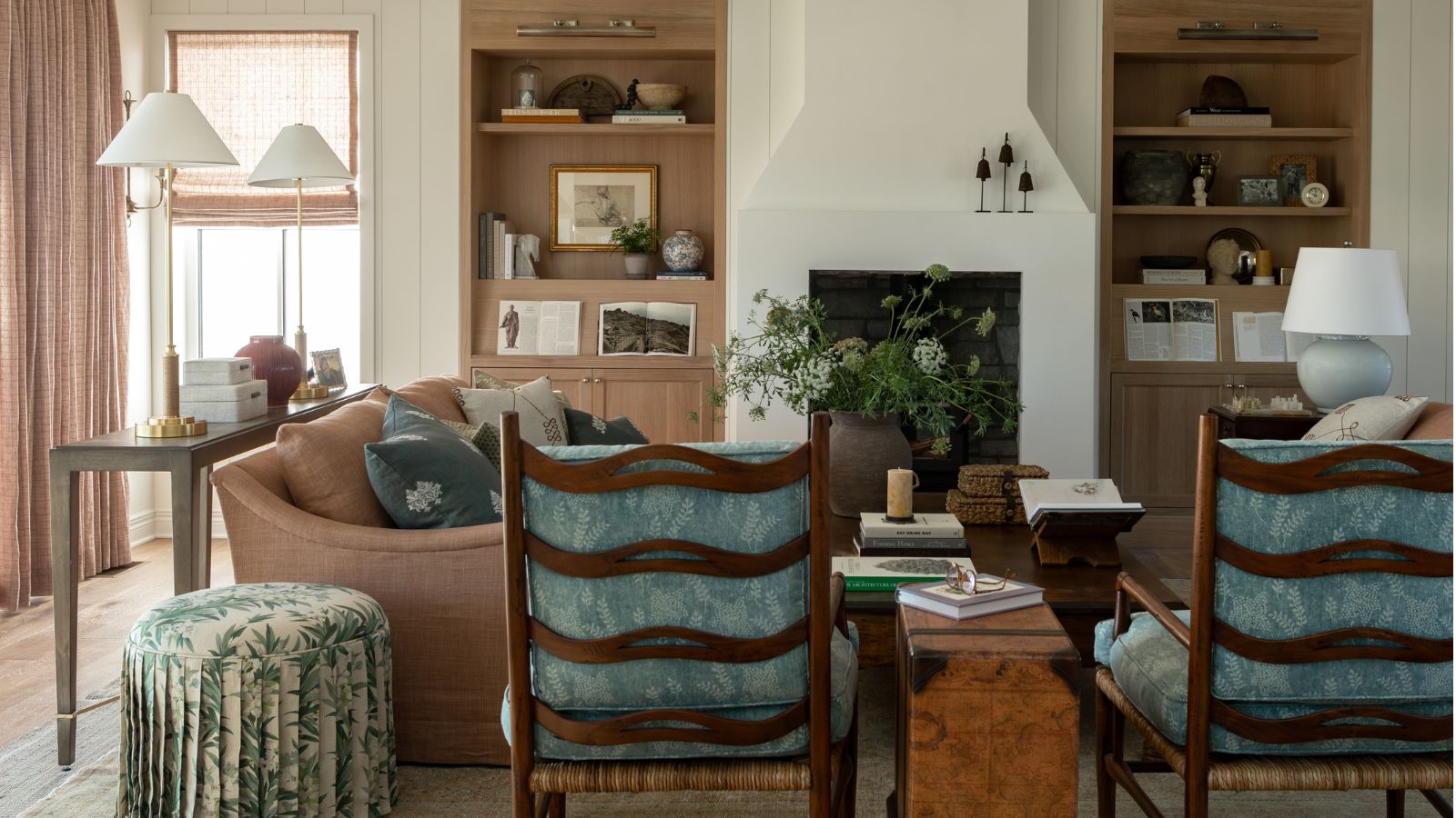 Bauernhaus trifft auf Oma-Chic, ästhetisches weißes Wohnzimmer mit Nut- und Federwänden, eingebauten Regalen aus heller Eiche, Kamin, zartrosa Couch, geblümtem Ottoman mit Rock und zwei Sesseln aus dunklem Holz mit blauen Kissen