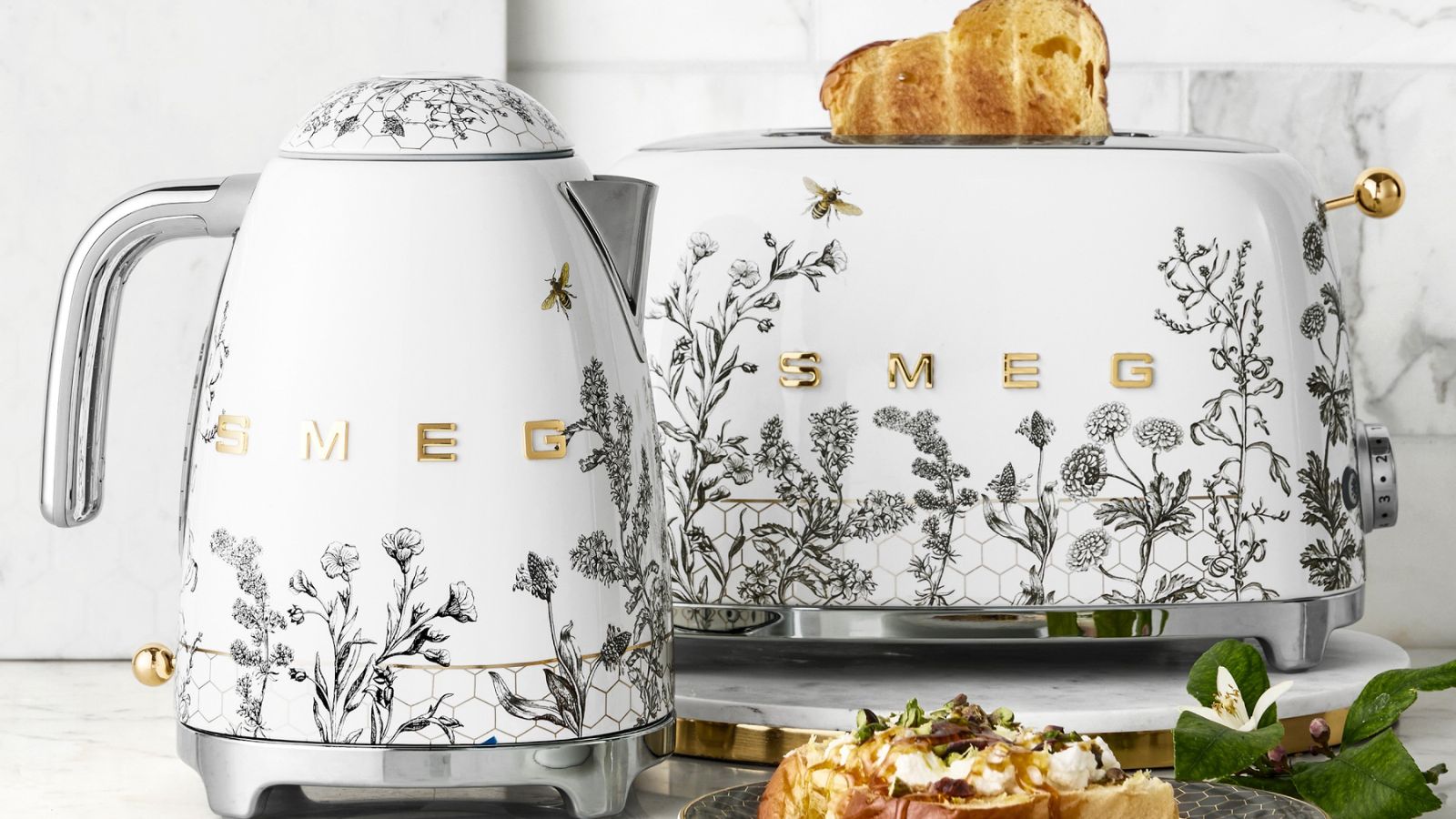 Ein SMEG-Wasserkocher und Toaster mit grauen Blumen- und Bienenmotiven auf weißem Sockel und Akzenten aus Edelstahl. 