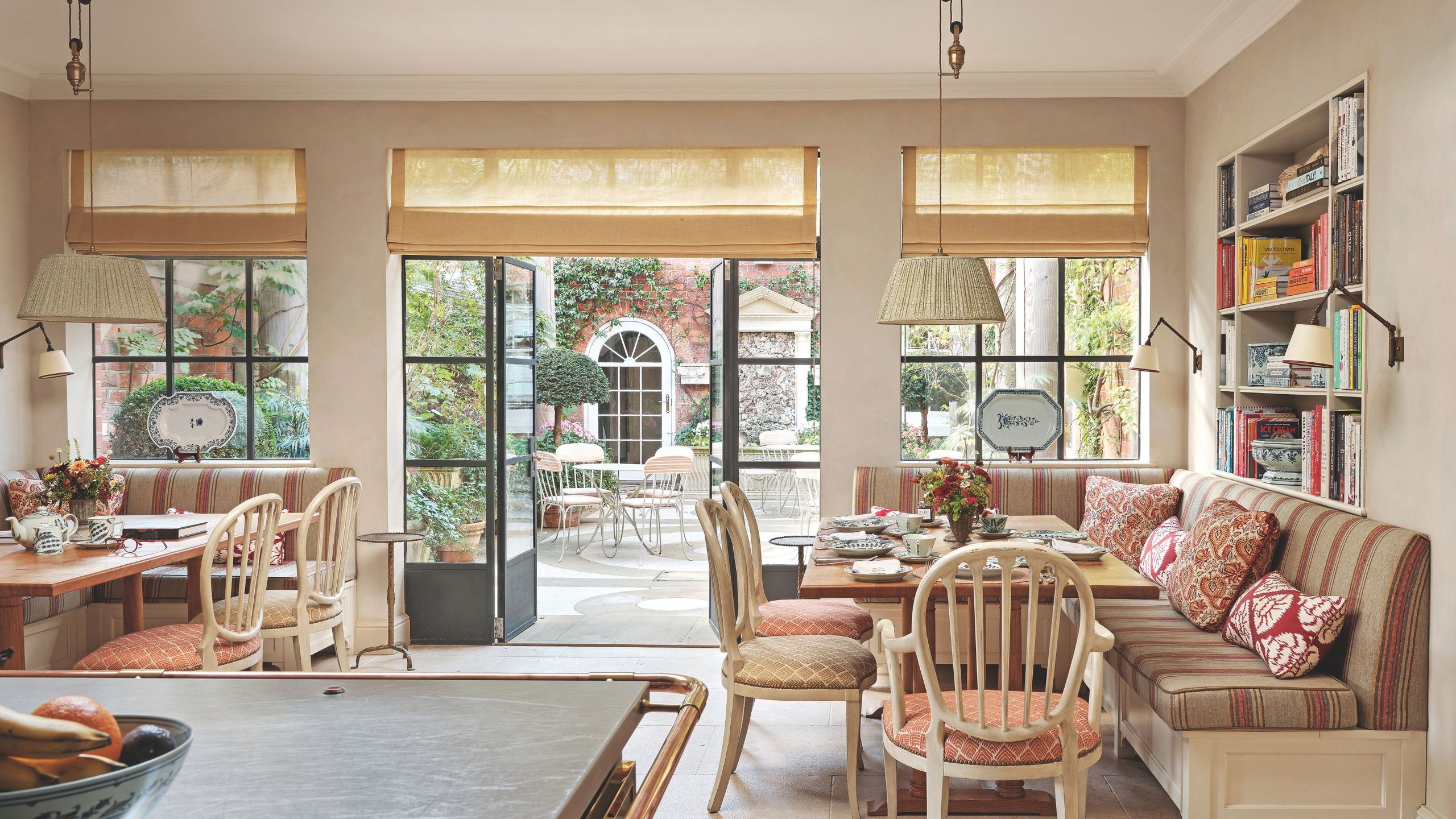 A bright breakfast nook with built-in banquette seating, patterned pillows, and large glass doors that open onto a lush garden courtyard