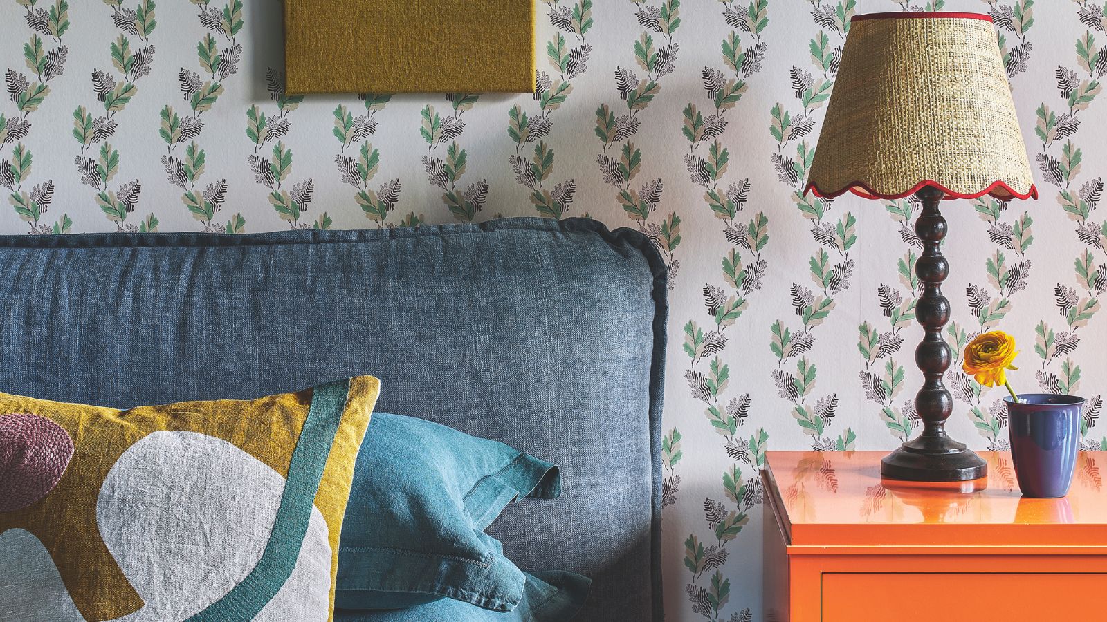 Blaues Kopfteil mit gemusterten Kissen, daneben ein orangefarbener Nachttisch mit Lampe mit Rattan, Wellenschirm und blauer Vase mit einer Blume. Dahinter befindet sich eine Tapete mit Blumenmuster.