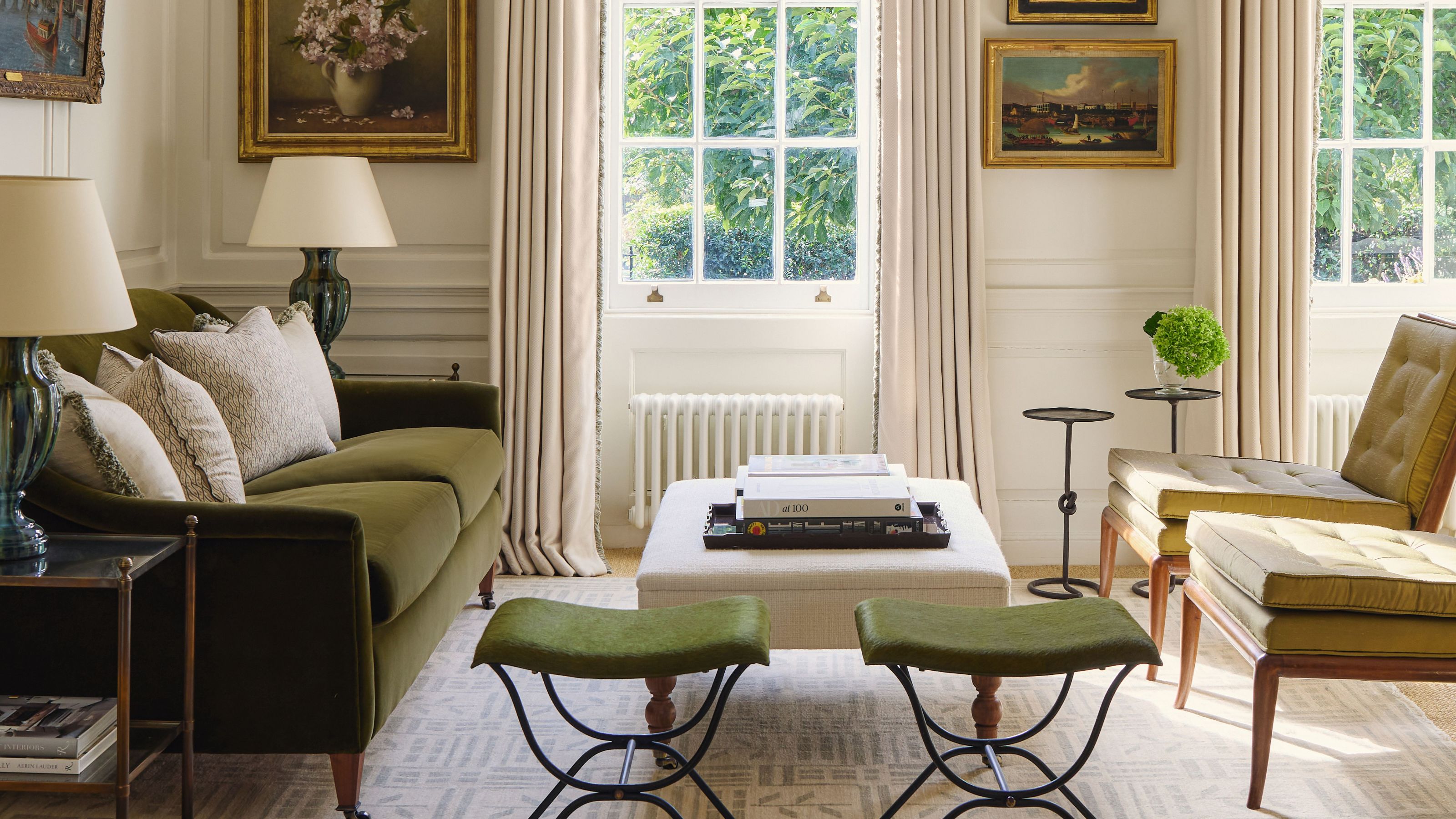 Ein helles, traditionelles Wohnzimmer mit einem waldgrünen Samtsofa, olivgrünen Mid-Century-Stühlen und zwei grünen Hockern. Große Schiebefenster sind von cremefarbenen Vorhängen und einem formellen Querbehang eingerahmt, an den Wänden hängen klassische Ölgemälde.
