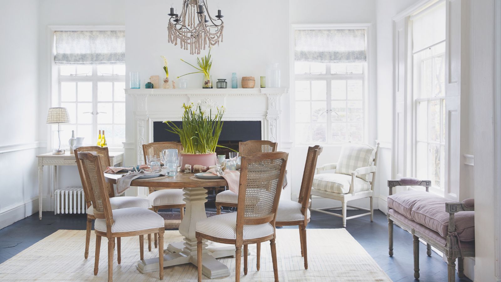 Ein helles, frühlingshaftes Esszimmer mit einem runden Esstisch aus Holz und Rattanstühlen in der Mitte sowie großen, hellen Fenstern rundherum mit transparenten Vorhängen. Hinten in der Mitte des Raumes steht ein großer weißer Kaminsims mit Blumen und Vasen, und der Tisch in der Mitte ist mit Narzissen, Tellern und Gläsern gedeckt. Unter dem Tisch liegt ein großer cremefarbener Teppich und rechts eine hellviolette Polsterbank.