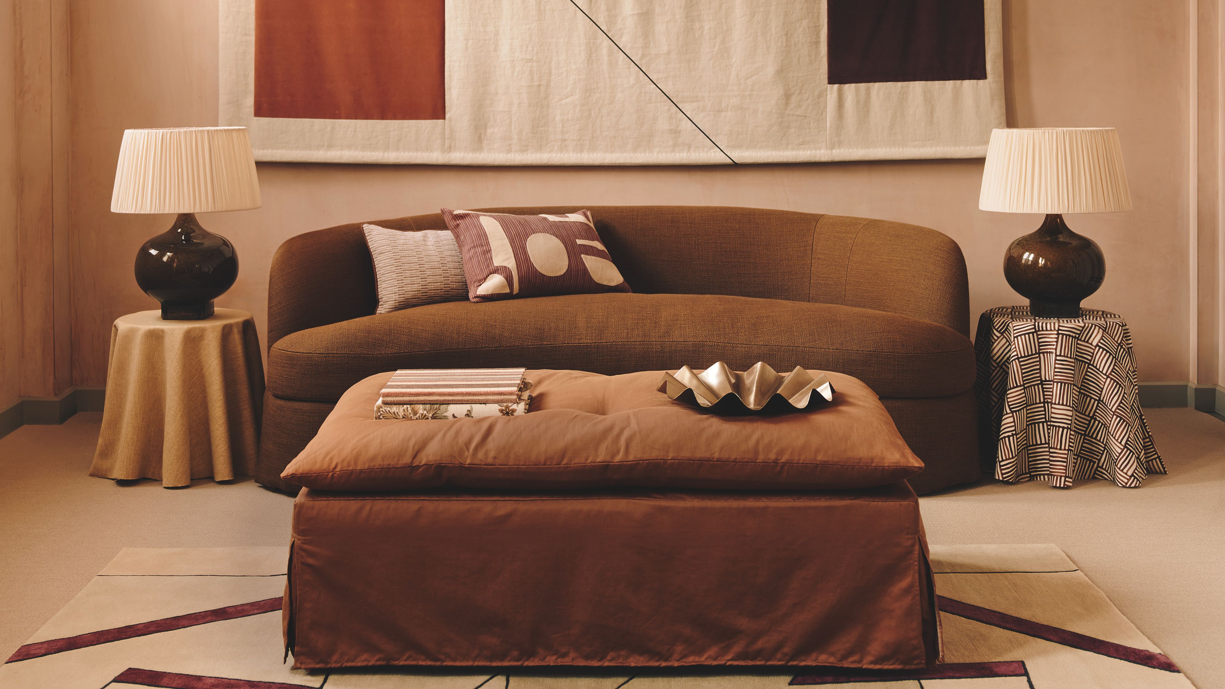 Ein warmes, terrakottafarbenes Wohnzimmer mit einem geschwungenen schokoladenbraunen Sofa und einem großen, abstrakten geometrischen Wandbehang. In der Mitte steht ein rostfarbener Ottoman auf einem gemusterten Teppich, flankiert von zwei Tischlampen mit plissierten Schirmen.
