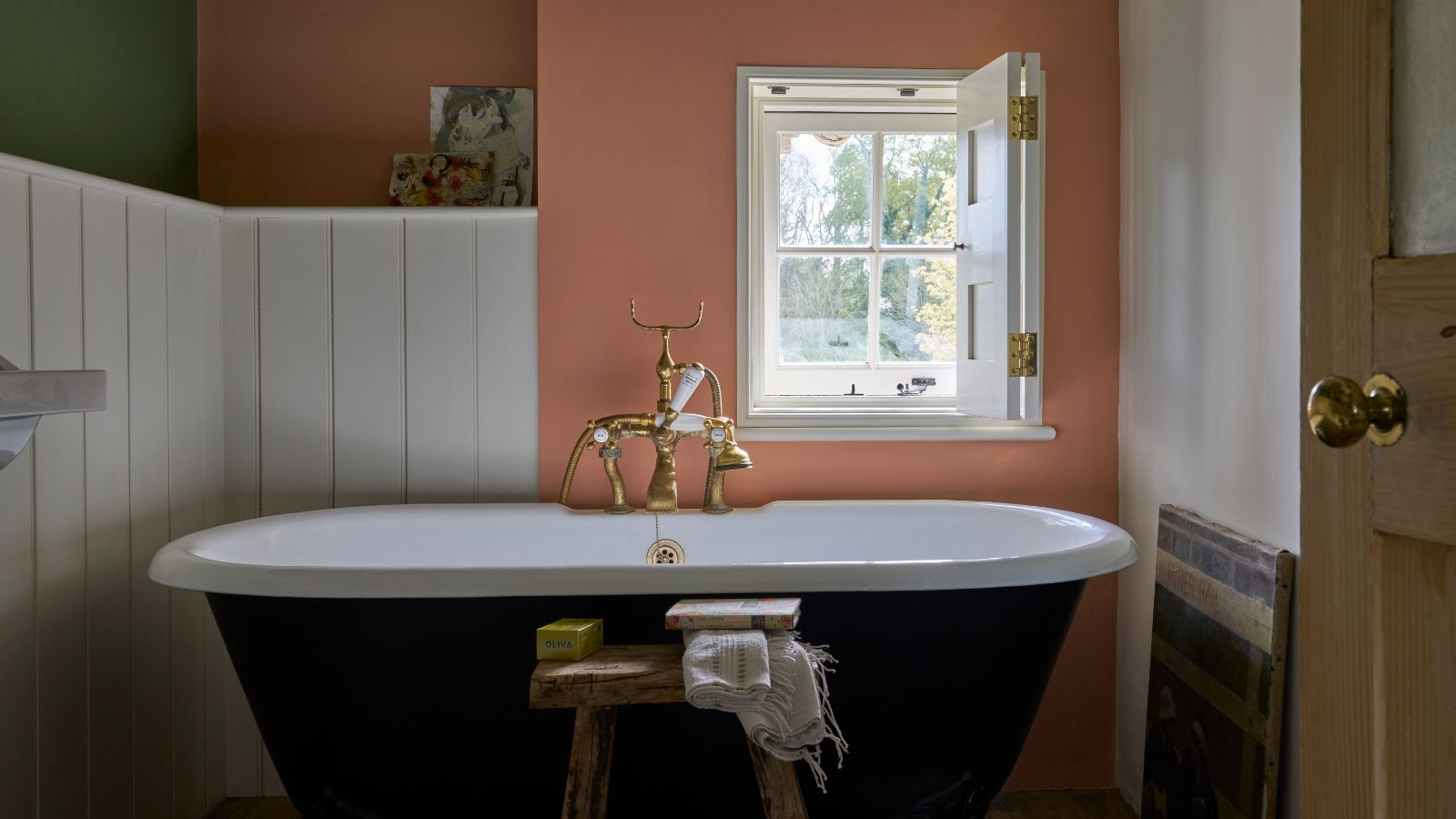 Ein Cottage-Badezimmer mit Terrakottawänden, einer freistehenden Badewanne, weiß getäfelten Wänden und einem kleinen Fenster. 