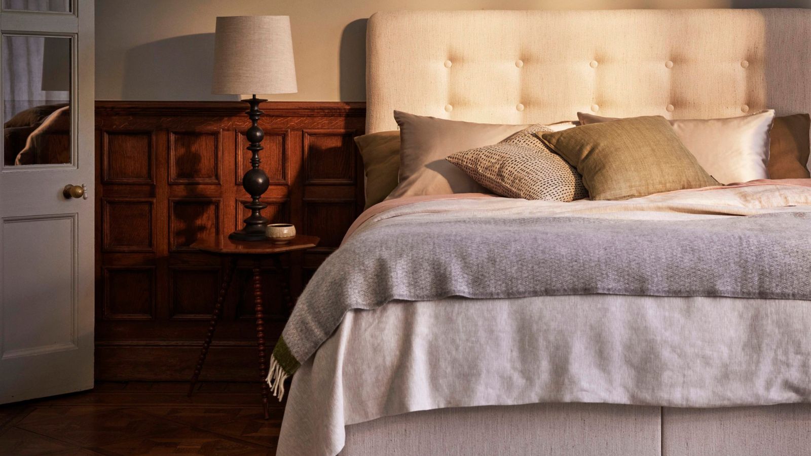 Neutraler Schlafraum mit einem cremefarbenen Polsterbett auf der rechten Seite, neutraler Leinenbettwäsche und Holzvertäfelung hinter dem Bett. Rechts neben dem Bett steht ein kleiner runder hölzerner Nachttisch mit einer dunklen Holzlampe mit weißem Lampenschirm und einer weißen offenen Tür daneben.