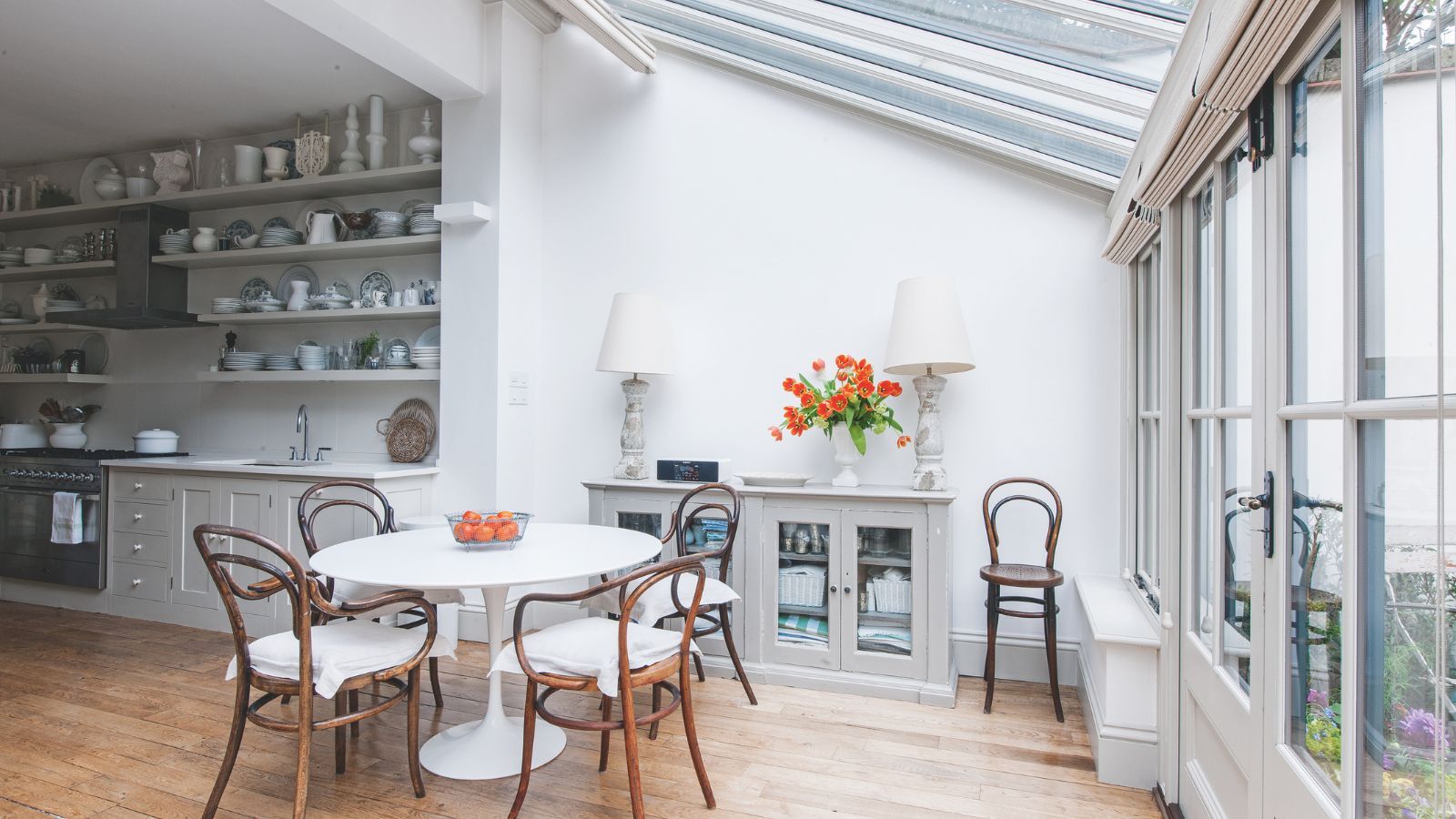 Conservatory with glass doors, round white breakfast table, wooden bistro chairs, a side console with two large lamps, leading into a kitchen with lots of open shelving decorated with ceramics