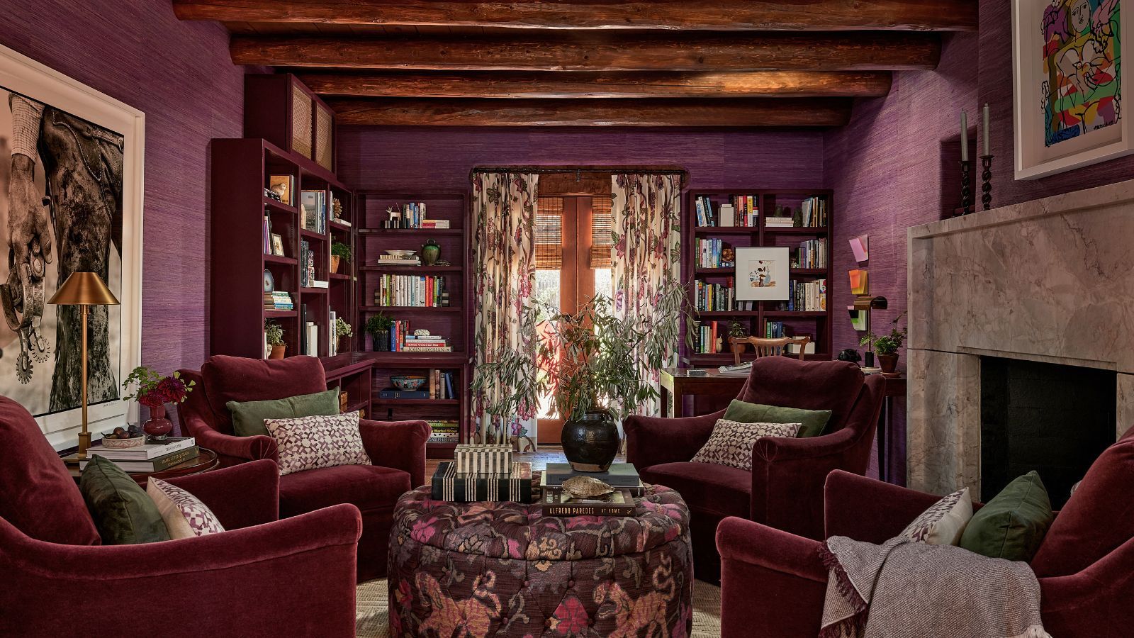 Living room with deep purple grasscloth walls, beamed ceiling, floral drapes covering glass doors, burgundy bookshelves, four maroon velvet armchairs sat around a floral ottoman with decor on top