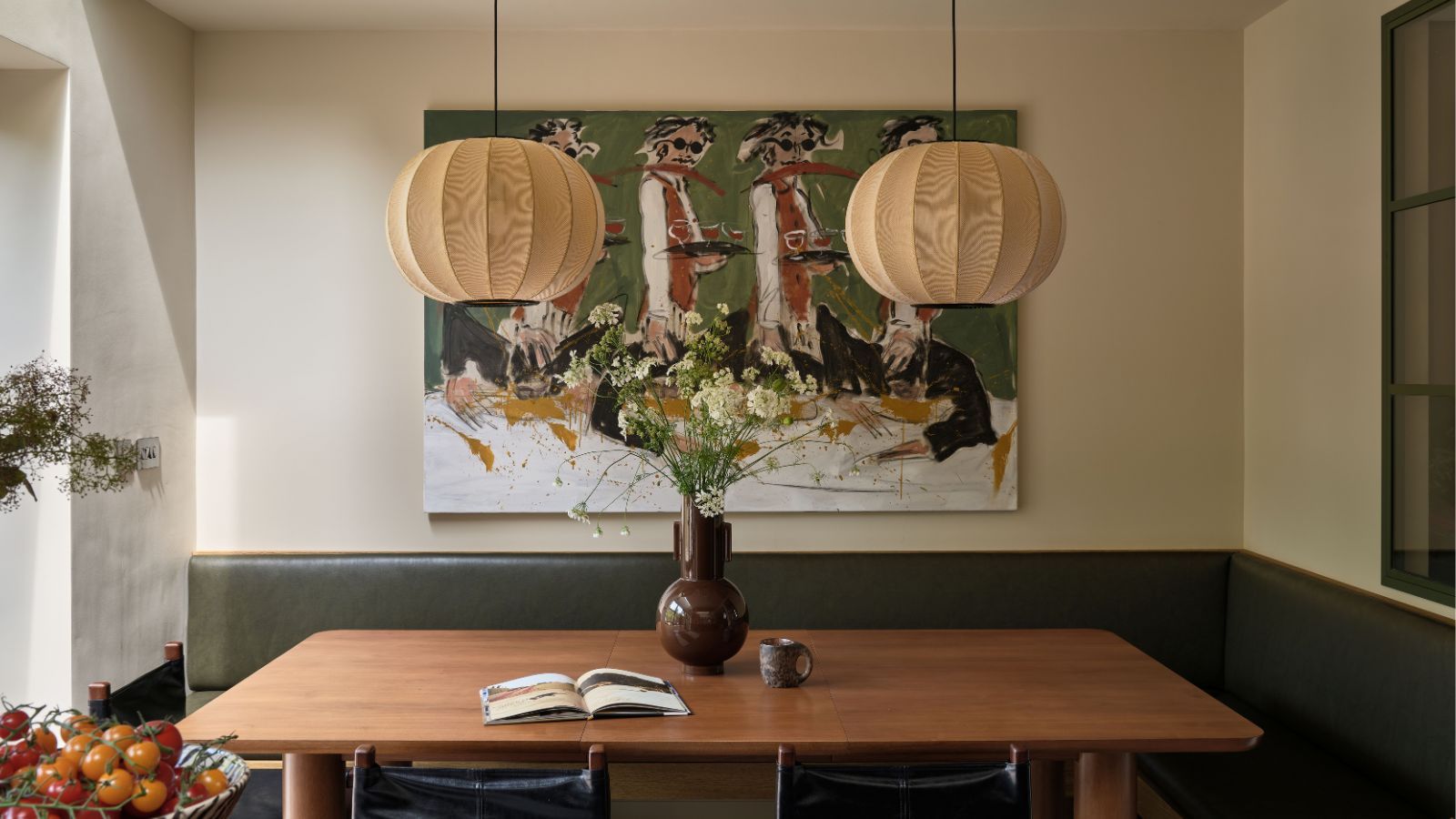 Dining nook with banquette green leather seating, a wooden table, a pair of lantern lights hanging above, an abstract painting of people dancing behind, and a large floral arrangement in a brown vase
