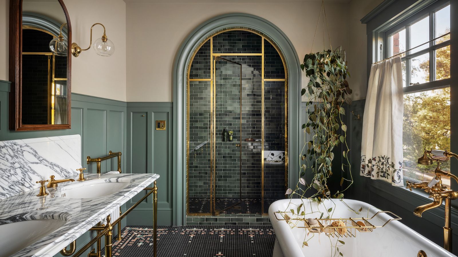 Ein elegantes Badezimmer mit gedämpfter grüner Wandverkleidung, cremefarbenen Wänden, einem Doppelwaschtisch aus Marmor mit Messingbeschlägen, einer geschlossenen Dusche mit gewölbter Tür und einer freistehenden Badewanne. 