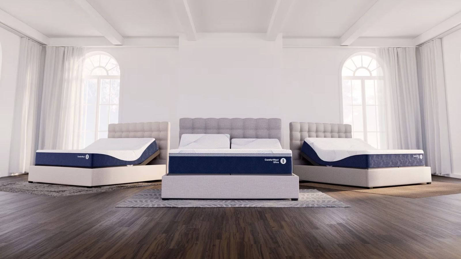 Drei Sleep Number-Matratzen auf grauen Betten in einem großen weißen Schlafzimmer mit dunklen Holzböden und zwei großen Bogenfenstern mit transparenten Vorhängen auf beiden Seiten.