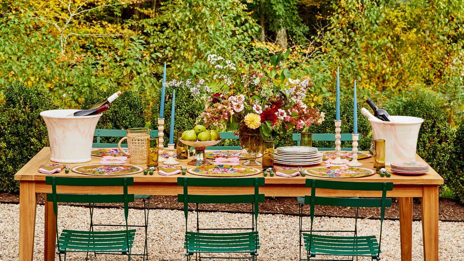 Sommerliche Tischlandschaft mit blauen Kerzen, großem Blumenarrangement, bunten Tischsets und Servietten, weißen Eiskübeln, alles auf einem hölzernen Esstisch mit grünen Eisenstühlen 
