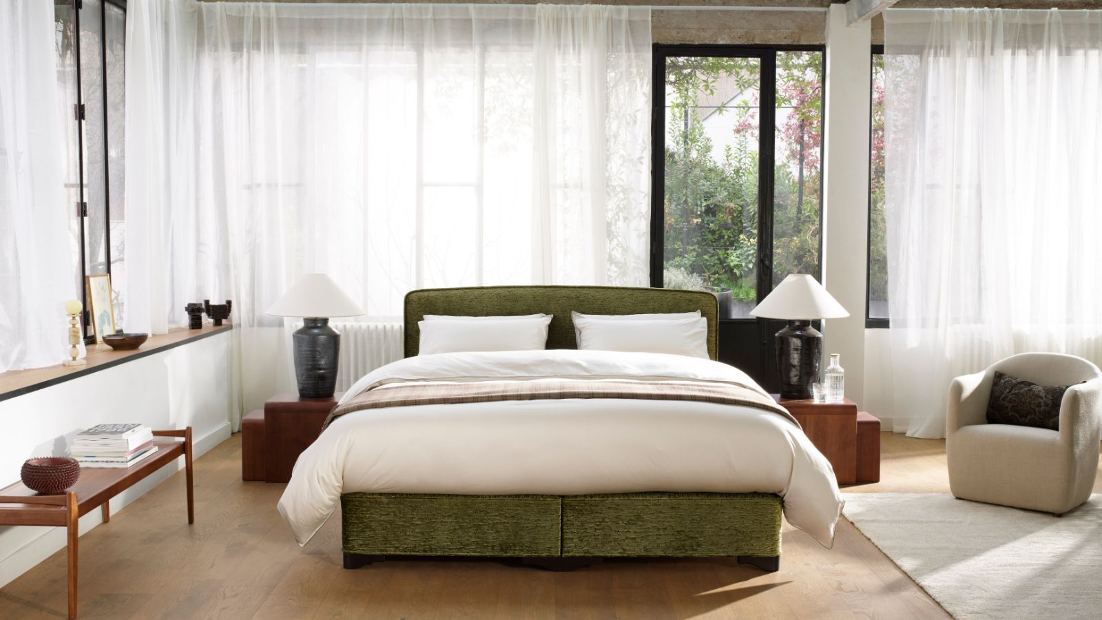 A large and bright bedroom with a green bed with white bedding, large wraparound windows with white sheer curtains, wooden flooring, a large white rug, and a white upholstered chair.
