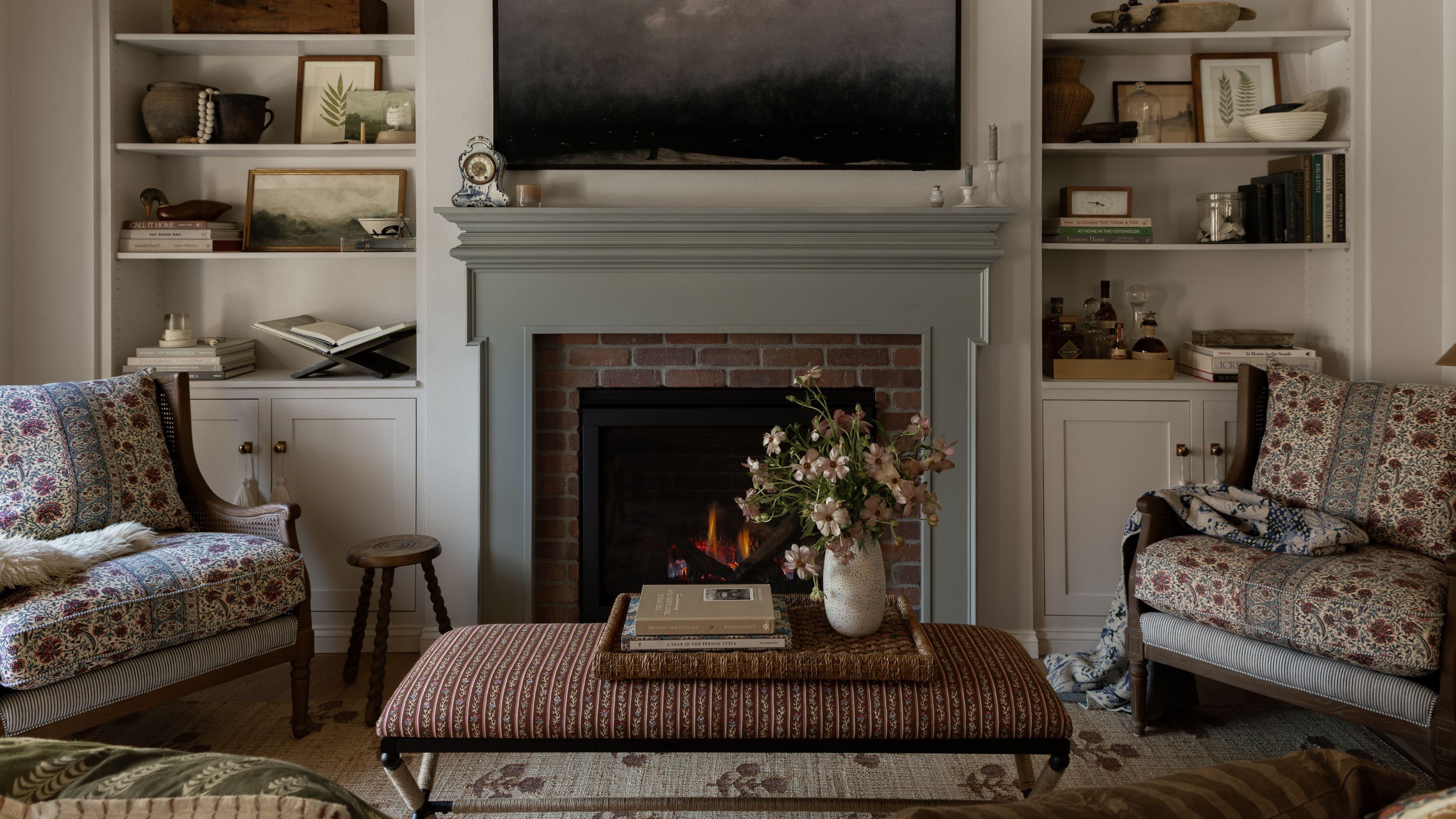 Ein neutrales kleines Wohnzimmer in einem Kutschenhaus mit einem sanftgrünen Kamin mit freiliegenden Ziegelsteindetails, eingebauten Bücherregalen, geblümten Sesseln, einem gestreiften Ottoman und einem gewebten Teppich mit floralen Details