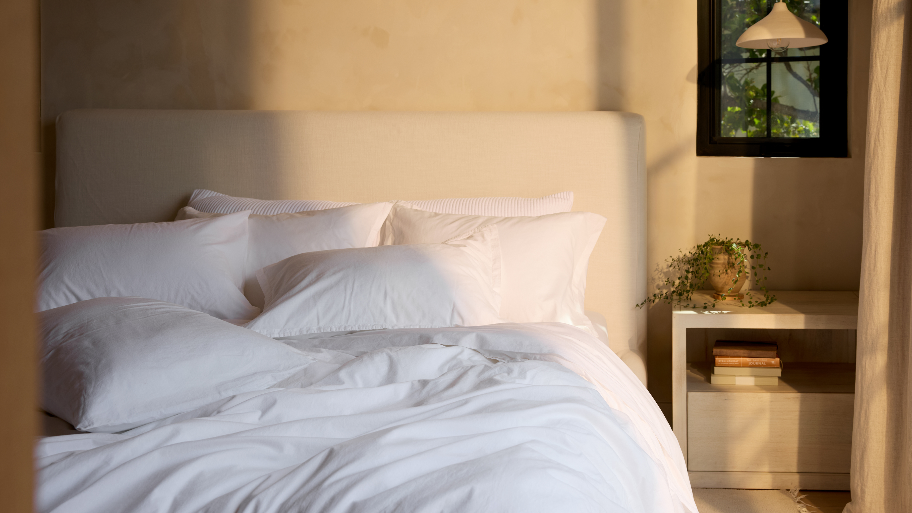 Neutrales Schlafzimmer mit minimalistischem Kopfteil und strahlend weißer Bettwäsche neben einem beigen Nachttisch und einer Pendelleuchte