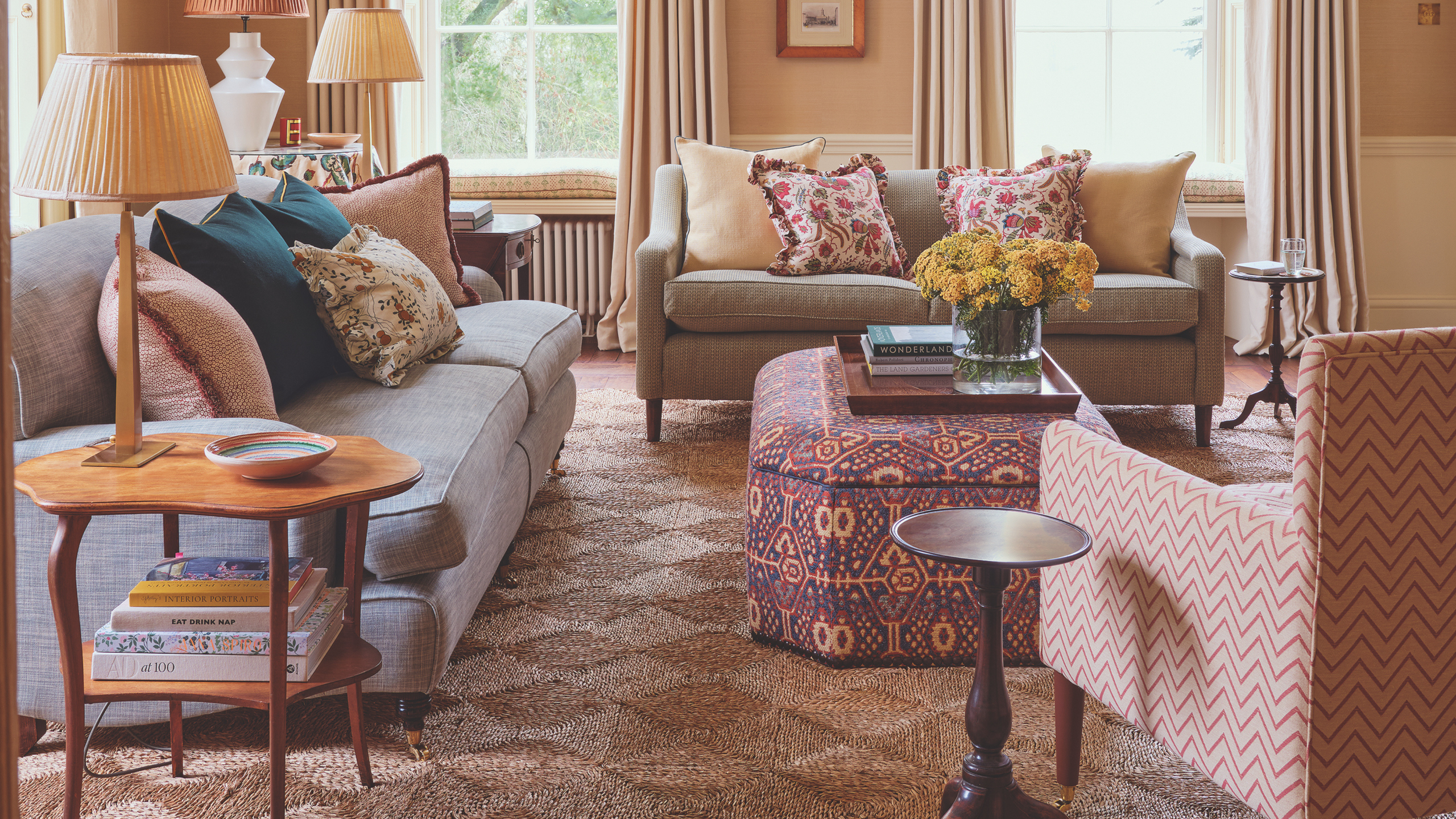 The cozy, pattern-filled living room of a 1700s country house restoration by Olivine Design, featuring an upholstered ottoman styled as a coffee table, holding a wooden tray of various objects.