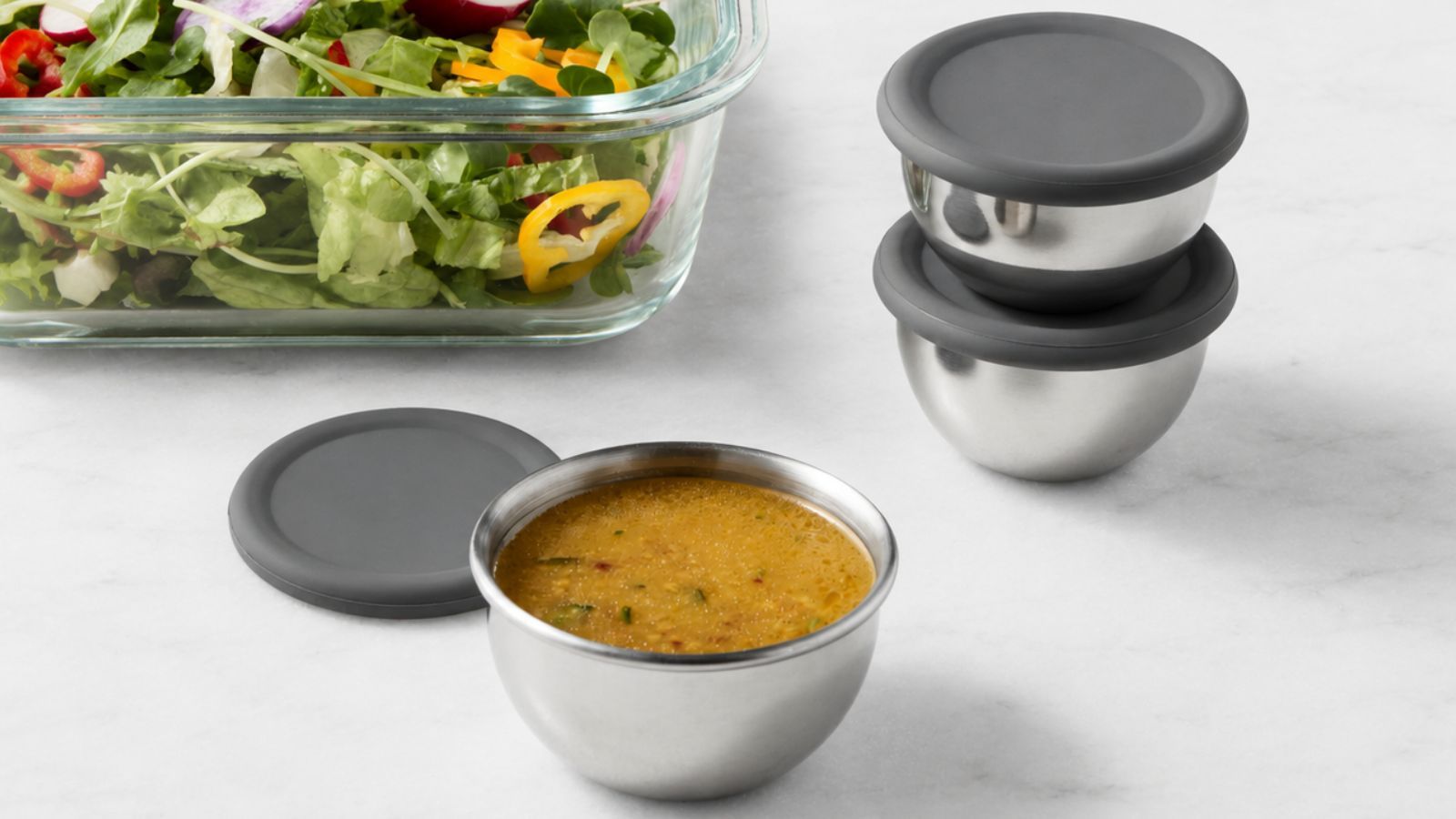 Three stainless steel condiment containers with grey silicone lids, on a white marble counter beside a glass dish of salad.