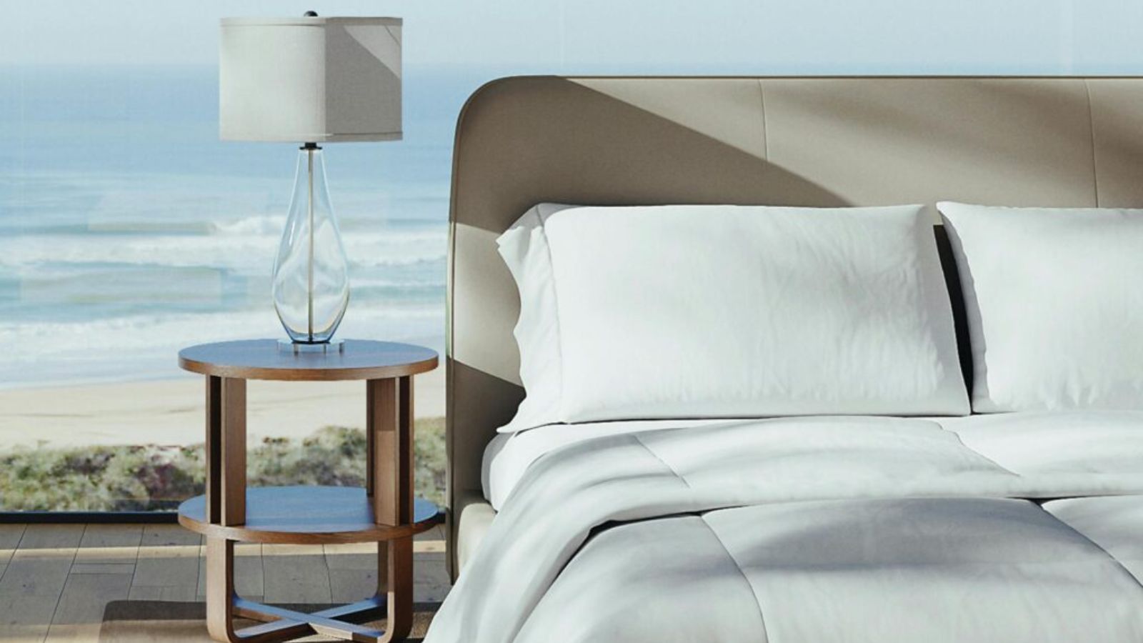 Ein Saatva-Bett mit weißer Bettwäsche neben einem Nachttisch aus Holz mit einer Glaslampe und einem weißen Lampenschirm. Dahinter liegt eine Strandlandschaft.