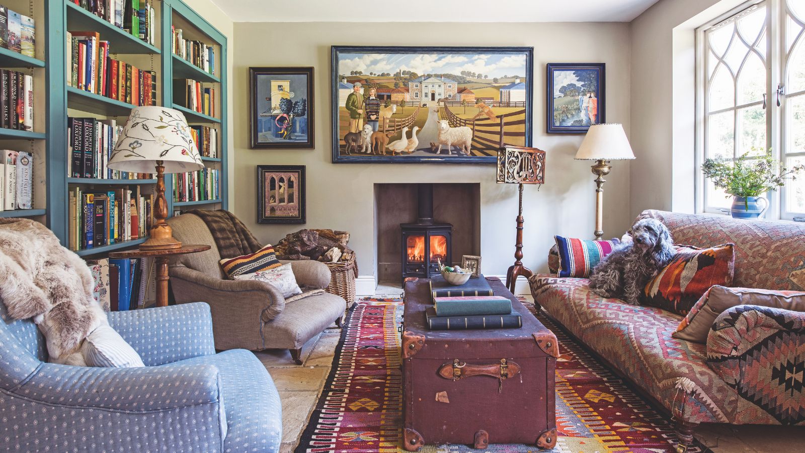 Ein gemütliches und vielseitiges Wohnzimmer mit einer Bibliothekswand, einem brennenden Holzofen, einem gemusterten großen Teppich, einer Truhe im Vintage-Look und einem nicht passenden gepolsterten Sofa und Stühlen.