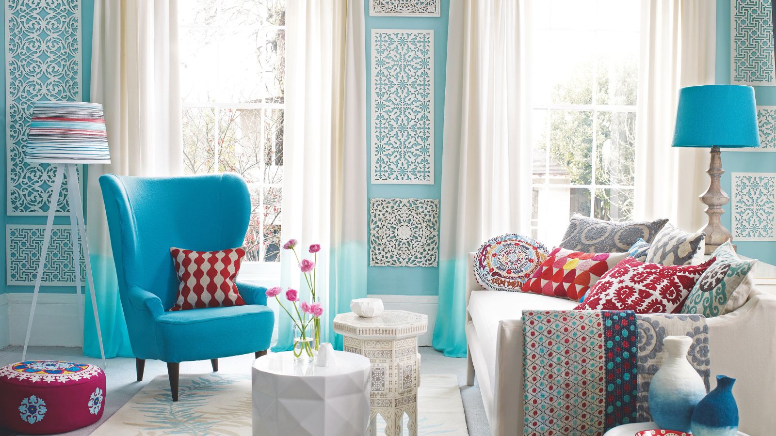 Helles und farbenfroh gemustertes Wohnzimmer mit blauen Wänden, einem blauen Stuhl, einer weißen Couch, einer blauen Lampe, einem weißen Couchtisch mit Blumen sowie gemusterten Decken, Kissen, Hocker, Tisch, Lampenschirm und Vasen.