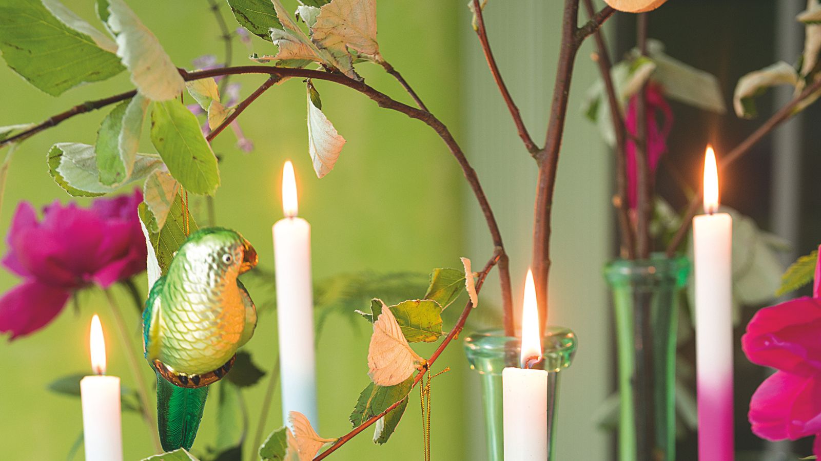 Brennende Stabkerzen vor einer hellgrünen Wand, abgebildet zwischen Zweigen und Grün mit rosa Blumen und einer grün glänzenden Vogeldekoration.