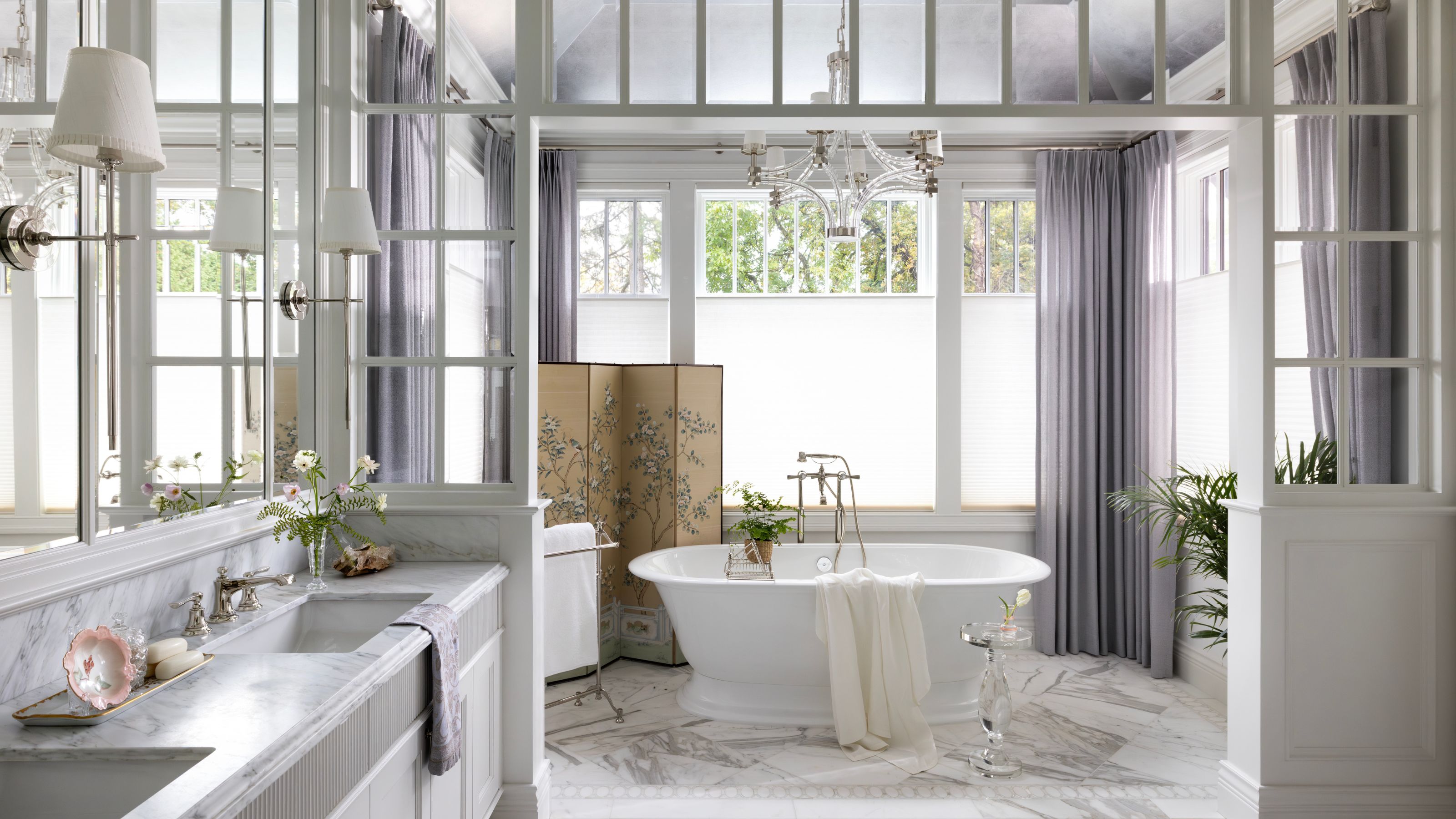 Helles Luxusbadezimmer mit einer weißen freistehenden Badewanne, Marmorböden, lavendelfarbenen Vorhängen und einem dekorativen Paravent unter einer Glasdecke.
